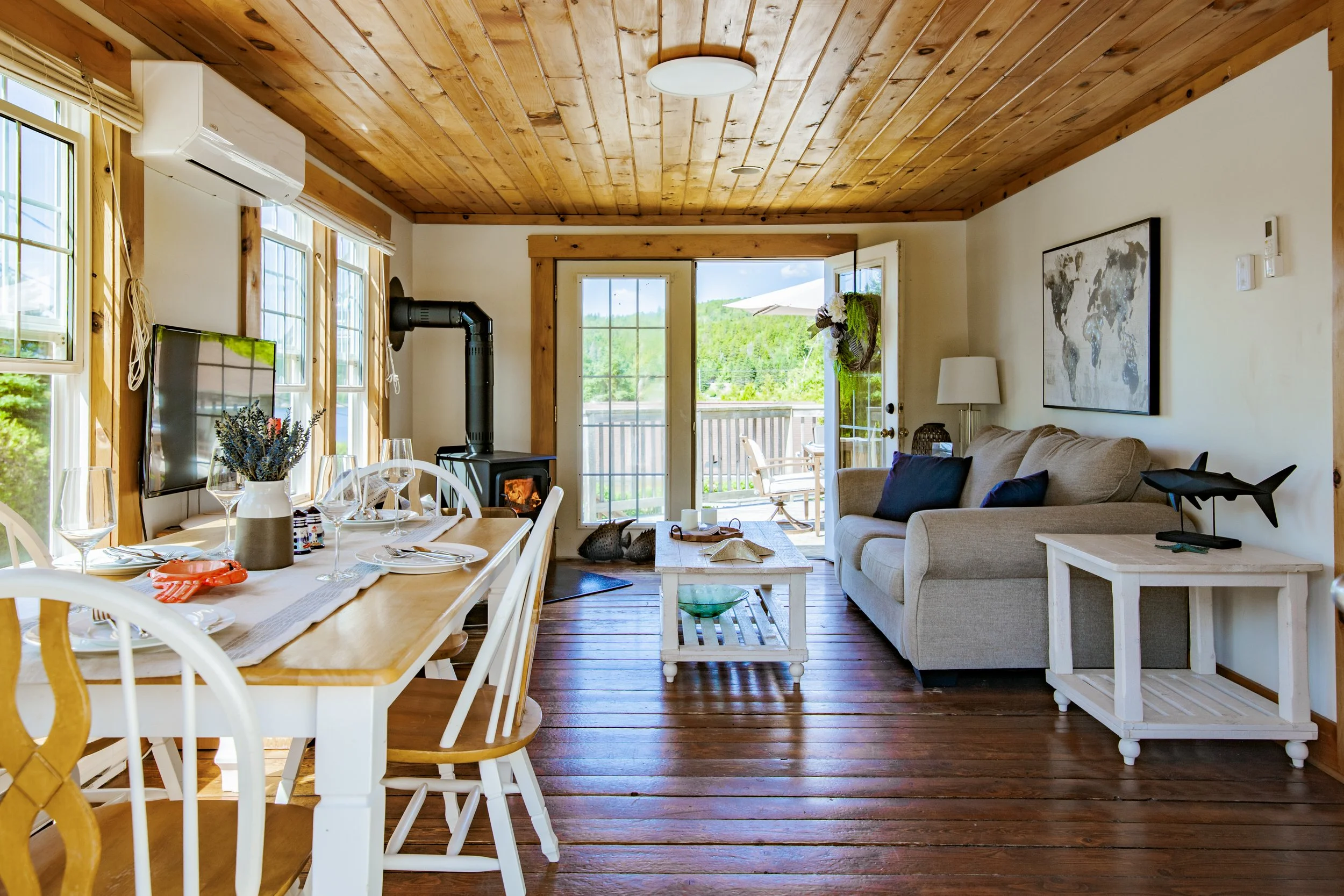 Living and dining rooms with wooden floors and ceiling, large windows, a door leading outside, a beige sofa, a white coffee table, a dining table set for six, a television, and a wood stove in a cozy, bright room.
