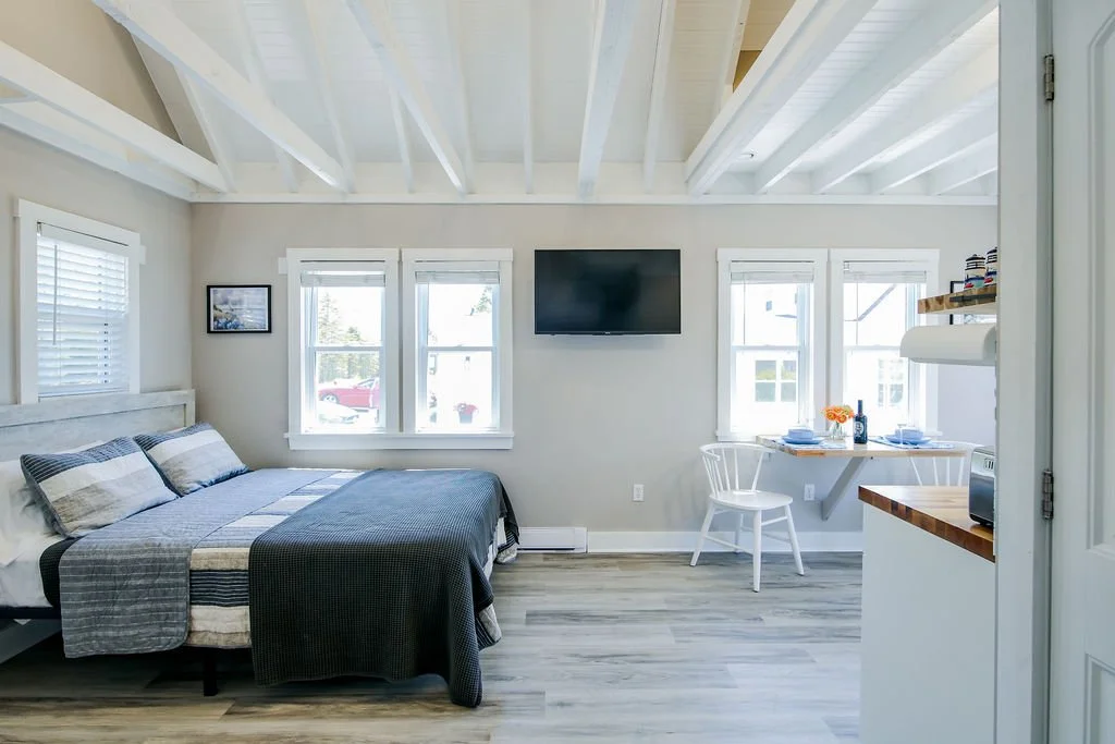 A bright, airy bedroom with a bed, three windows, a wall-mounted TV, a small table set for two, and light-colored wood flooring.