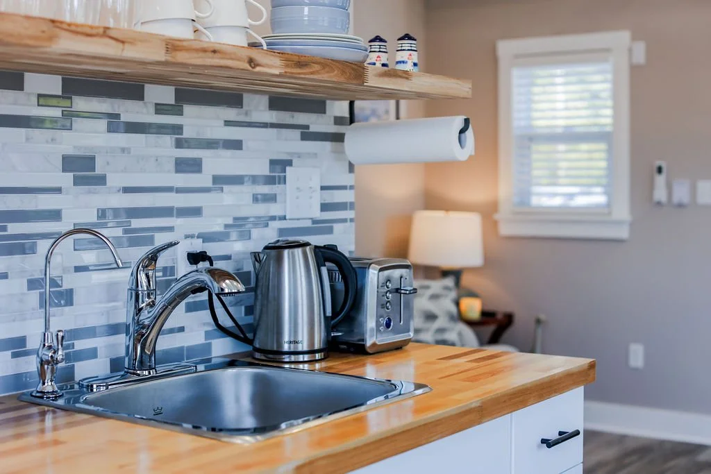 Kitchen sink with a chrome faucet, electric kettle, toaster, and paper towel holder on a wooden countertop, tiled backsplash, open shelf with white and blue dishes, and a living area with a couch, lamp, and window in the background.