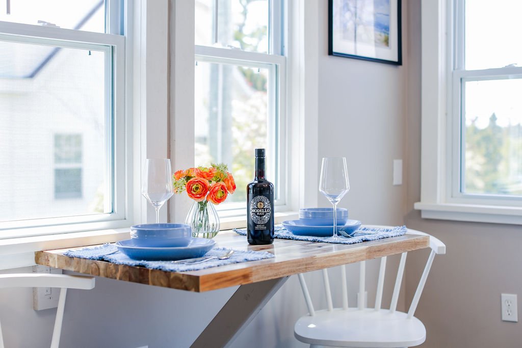 A dining table set for two with blue dishes, wine glasses, a bottle of wine, and a small bouquet of orange and yellow flowers in a glass vase, near two large windows in a bright room.