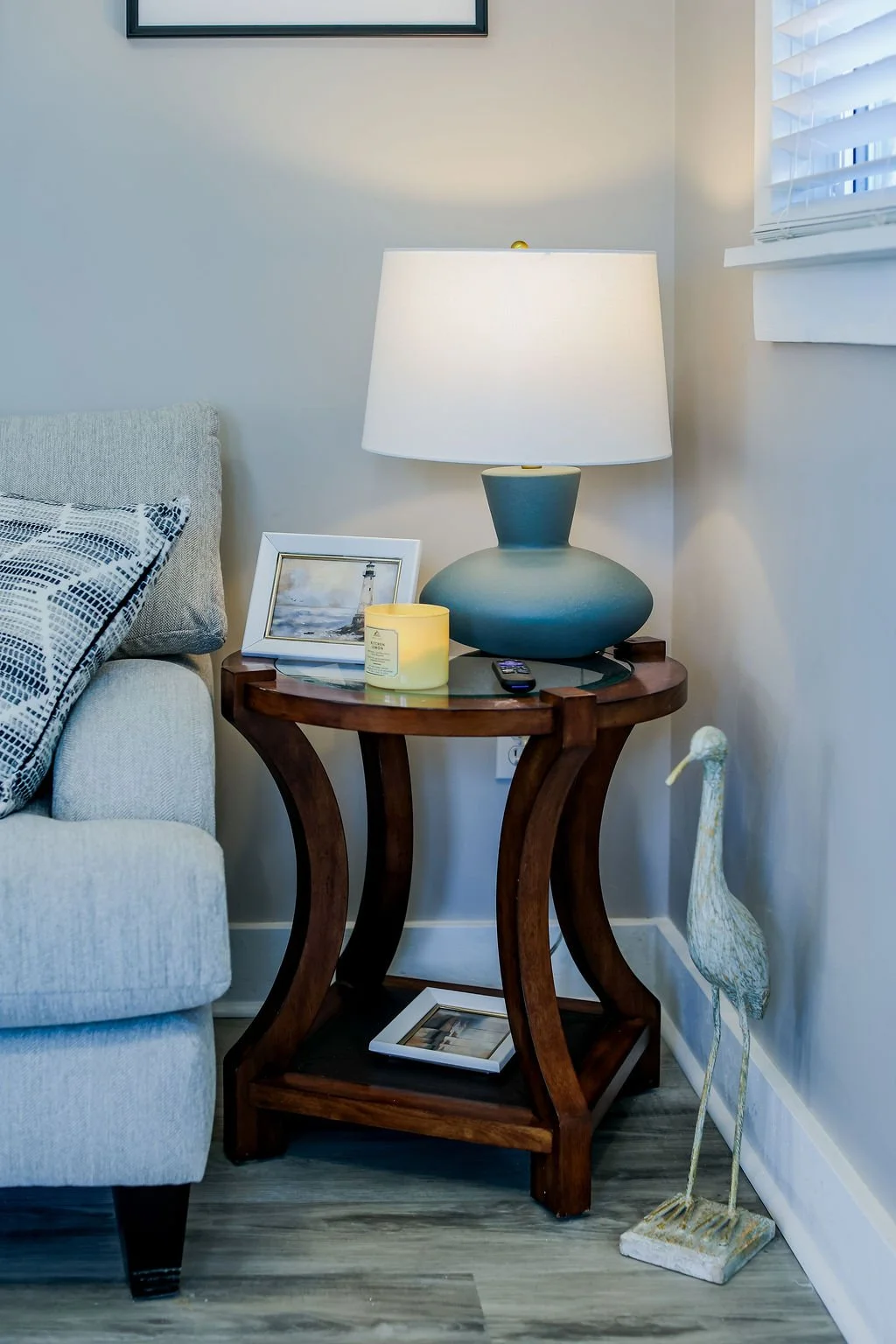 A living room corner with a beige couch, a wooden side table holding a lamp, a framed photo, a candle, and a remote, a pelican sculpture on the floor, and a window with white blinds.
