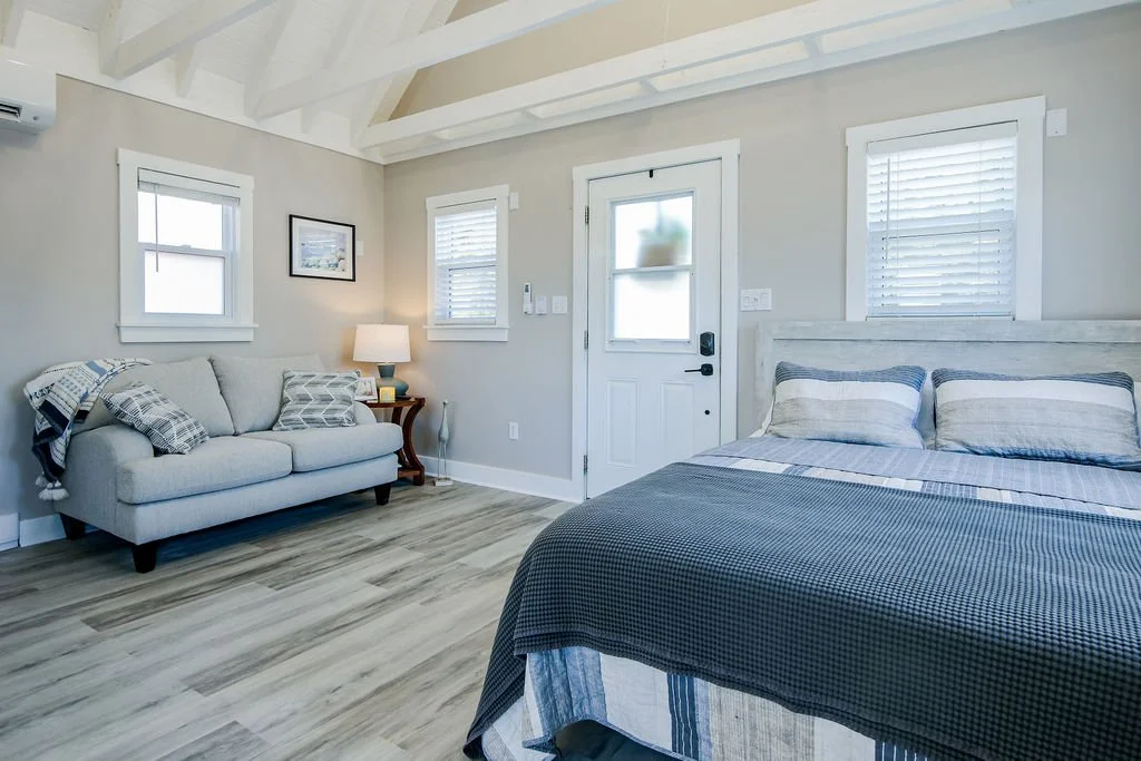 A bedroom with a gray bed, white walls, three windows with blinds, a gray sofa with pillows, a wooden side table with a lamp, and a door with a frosted window.