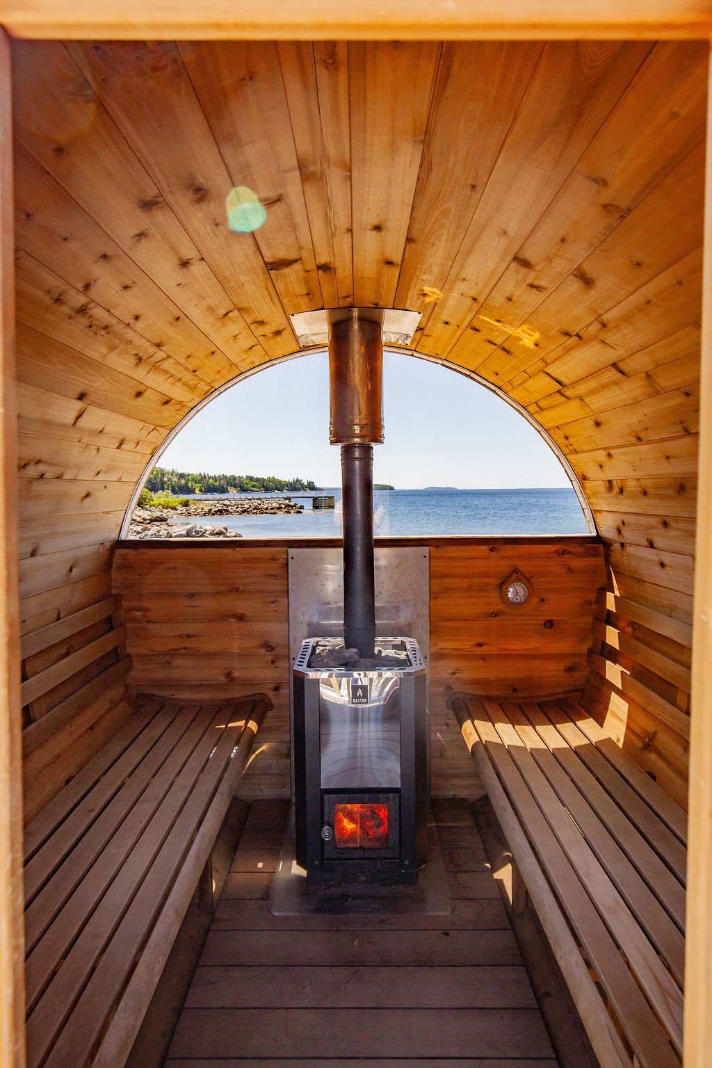 Interior of a wooden sauna with two benches, a stove, and a view of a body of water through an arched window.