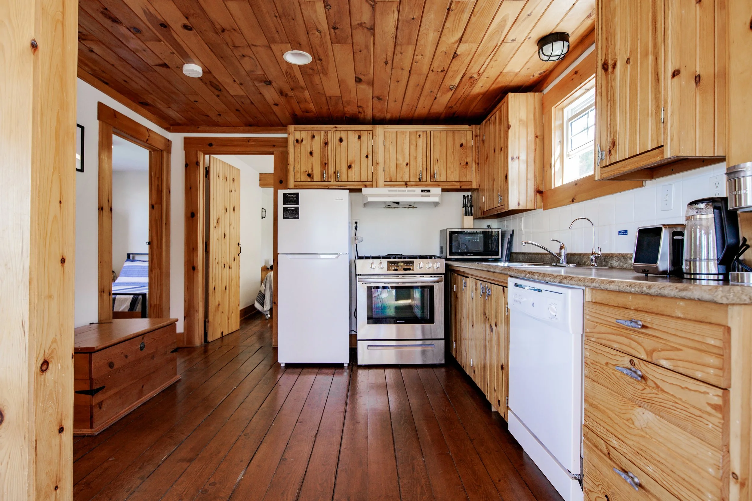 A kitchen with wooden cabinets, a fridge, a stove, microwave, dishwasher, and wooden flooring and ceiling. There's a small window above the sink.