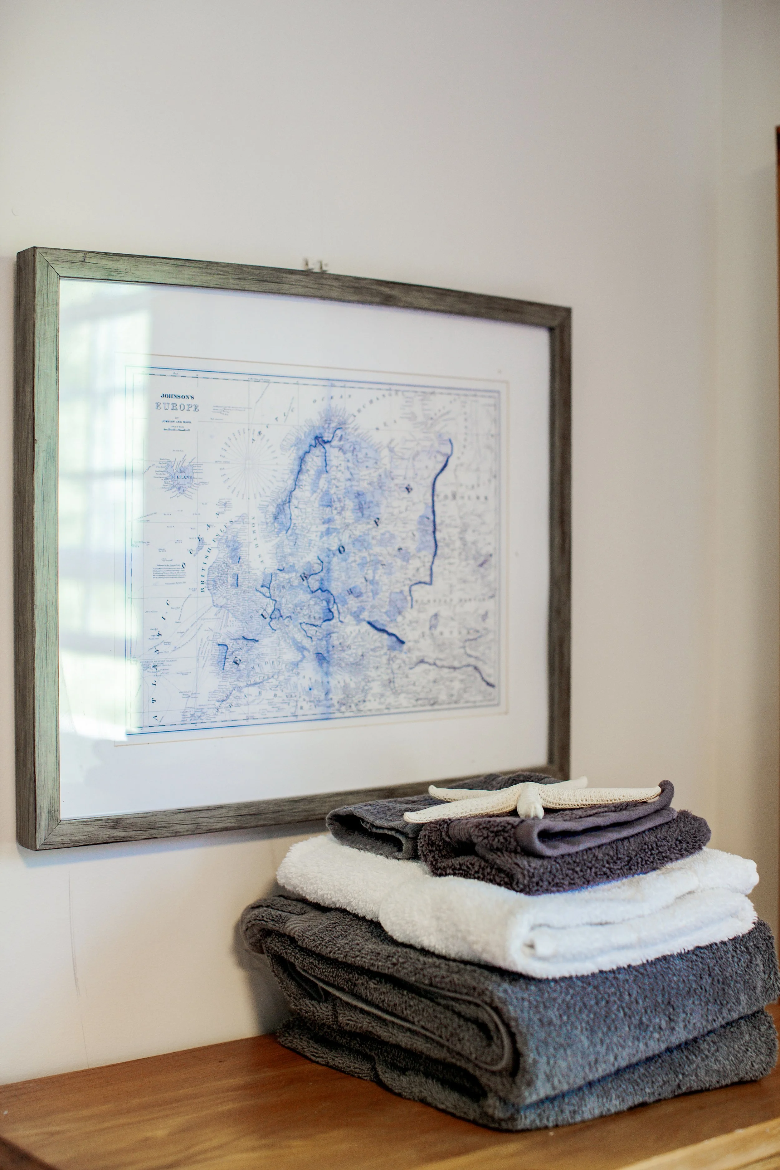 Stacked folded towels in white, dark gray, and light gray on a wooden surface beneath a framed map of Europe with a glass reflection in the background.