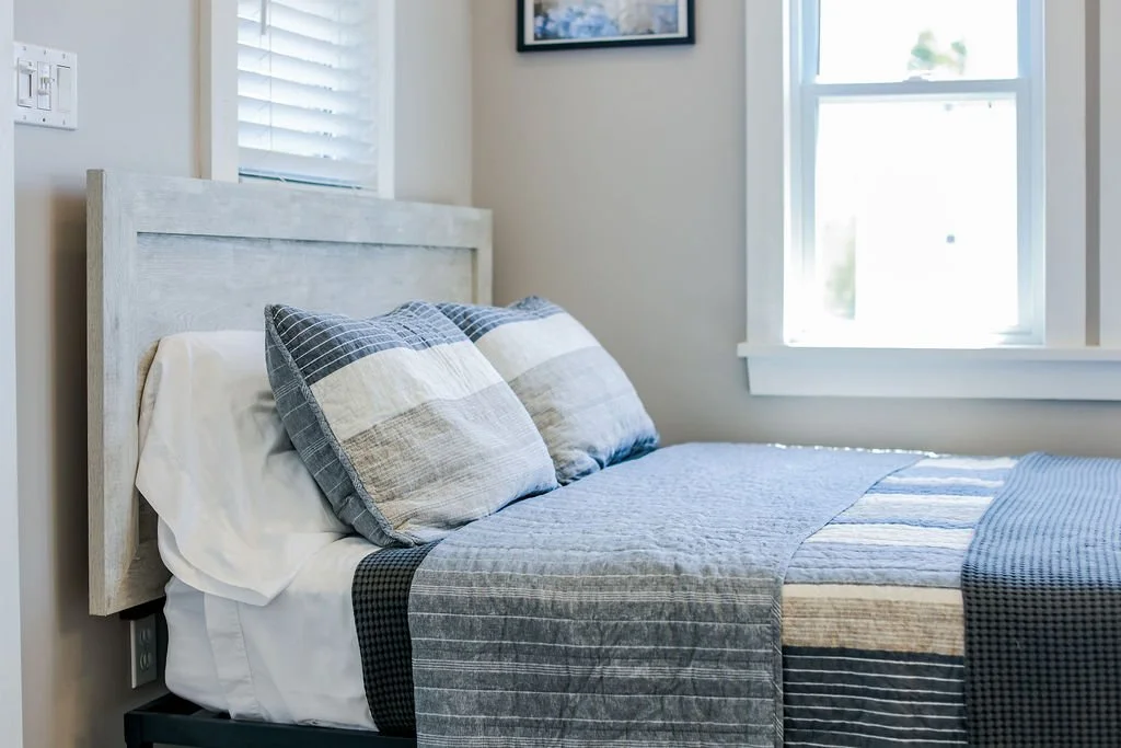 A neatly made bed with a light-colored wooden headboard, set next to a window with white blinds, featuring pillows with striped covers and a quilted bedspread.