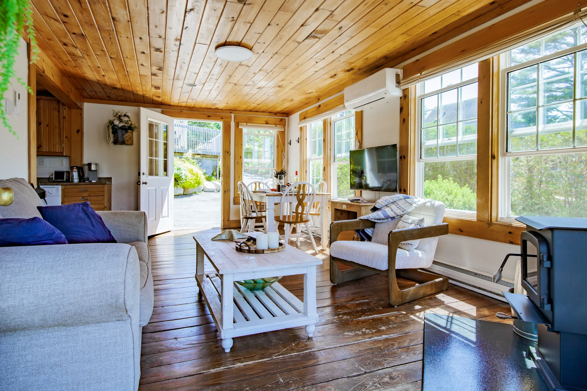 Cozy living room with wooden ceiling and floors, large windows letting in sunlight, a gray sofa with purple pillows, a white armchair with a blanket, a wooden coffee table, a TV on a wooden stand, and an open door leading outside to a garden.