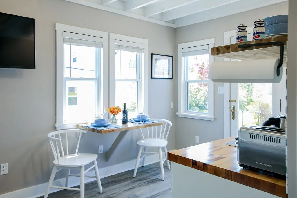 Bright kitchen corner with two windows, white chairs, a small wooden table set with blue plates and a wine bottle, a wall-mounted paper towel holder, and a toaster on a wooden countertop.