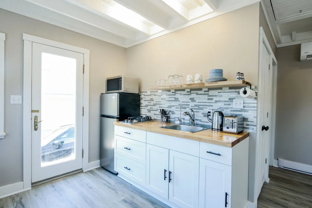 Kitchen with white cabinets, a wooden countertop, a small stainless steel refrigerator, microwave, toaster, electric kettle, and a dish rack. The backsplash is a gray-tile mosaic, and there's a door with a window leading outside.