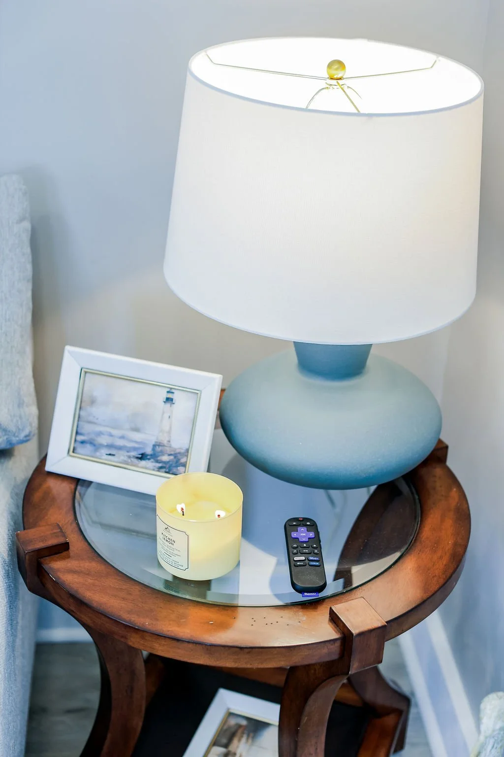 A bedside table with a table lamp, a framed picture of a lighthouse, a lit candle, and a remote control.
