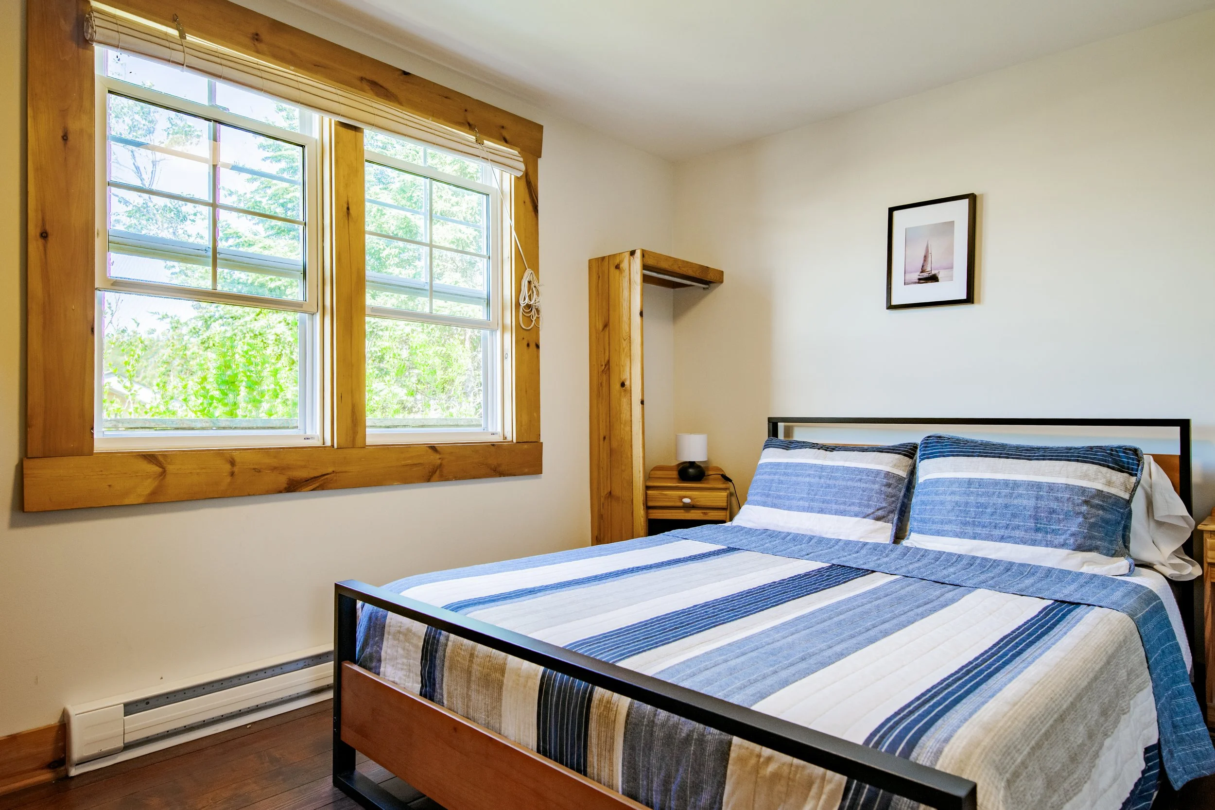 Bedroom with a bed covered in a blue and white striped quilt, two pillows, wooden window frames, a nightstand with a lamp, and a framed picture of a sailboat on the wall.