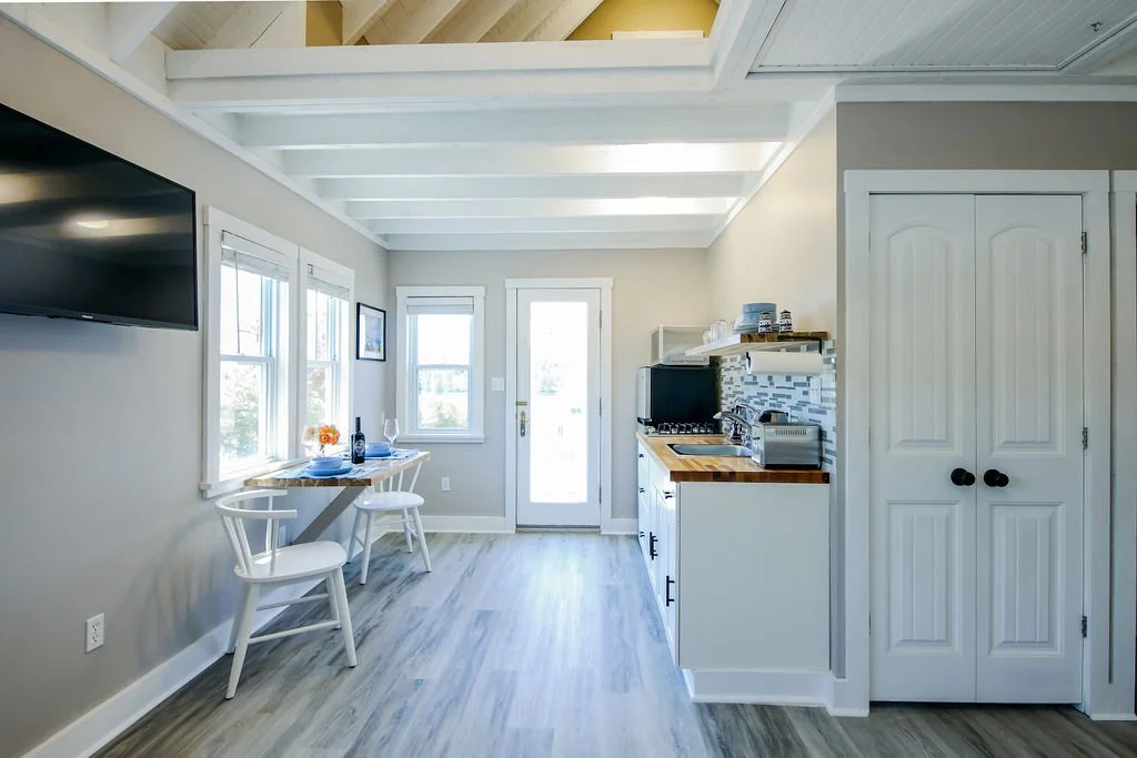 Bright, minimalistic kitchen and dining area with white walls, ceiling beams, small table with two chairs, windows, and kitchen appliances.