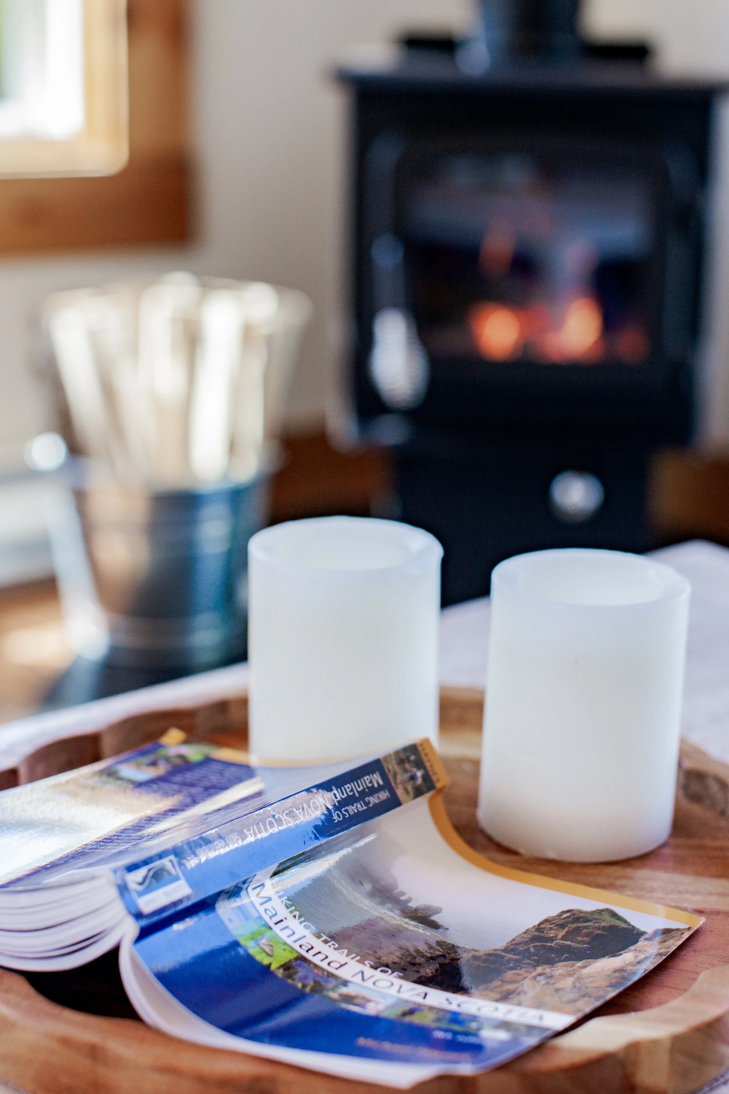 A wooden tray with camping brochures, two white candles, and a metal bucket holding wooden utensils, with a black fireplace in the background.