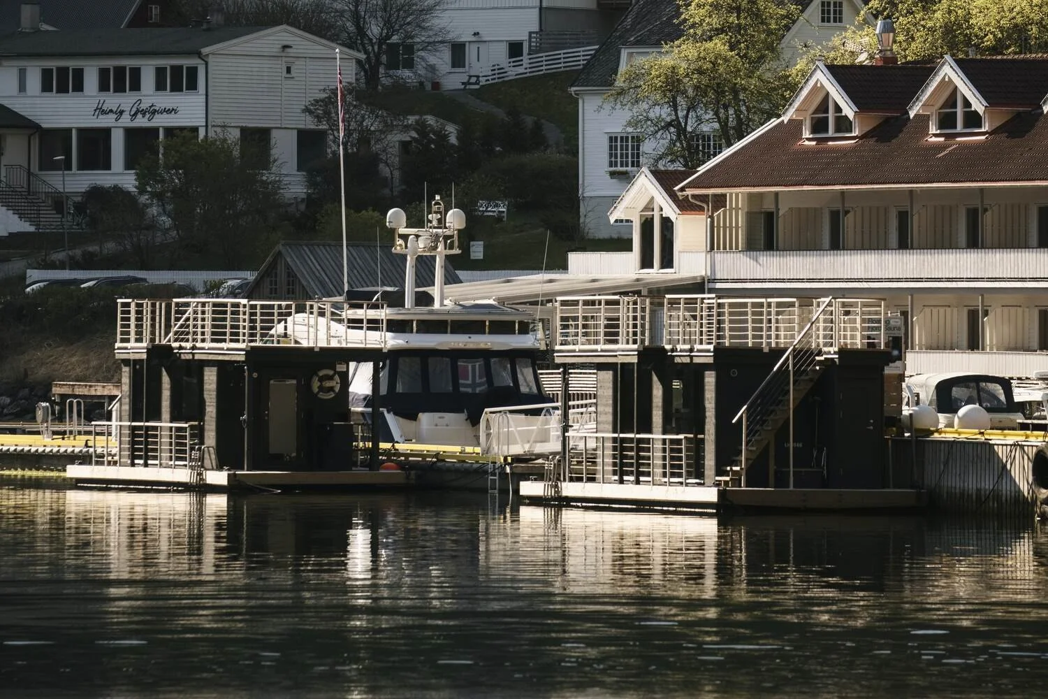 Floating saunas docked near a coastal building with trees and other houses in the background.