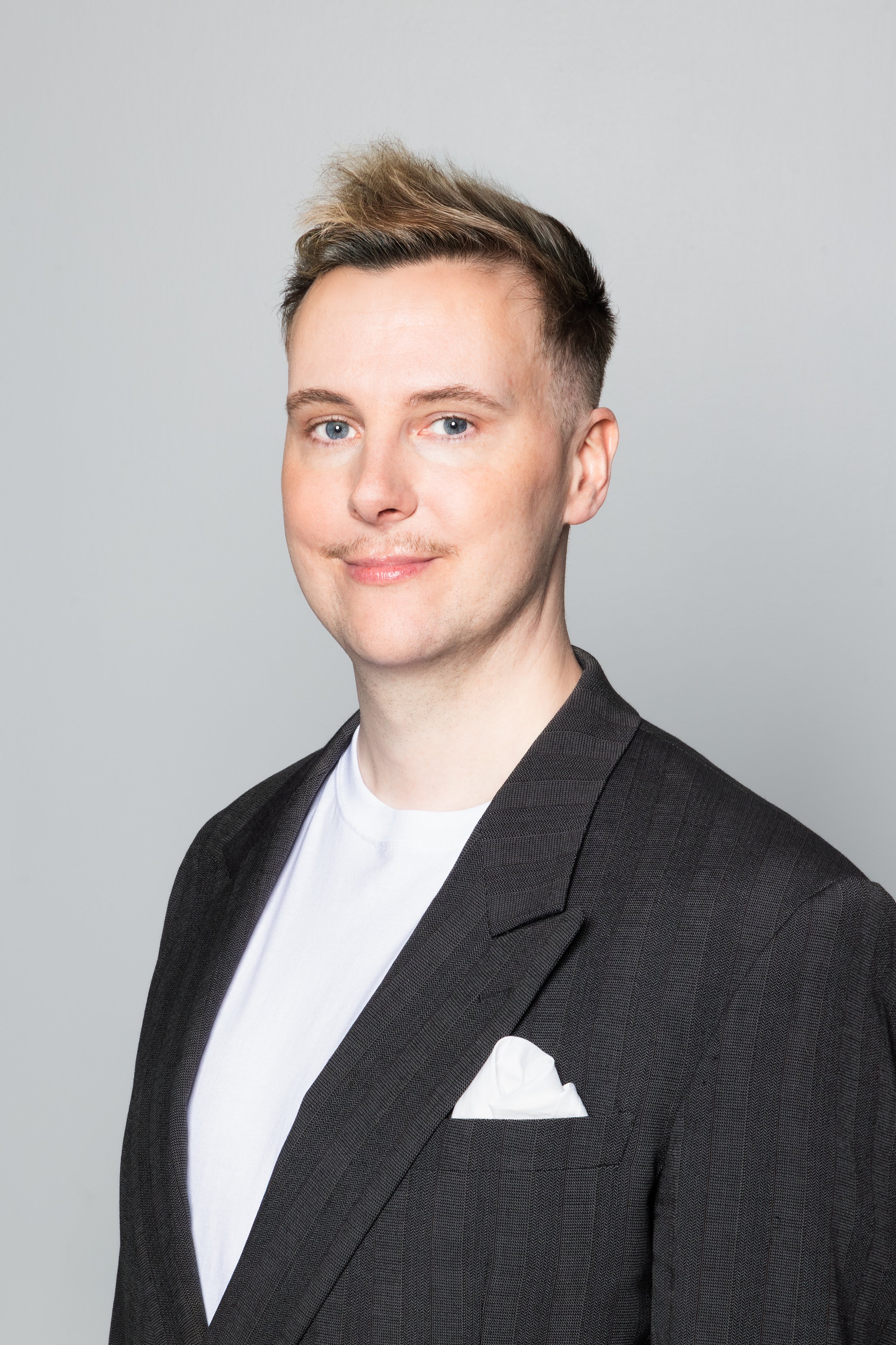 Guðni has short blond hair and blue eyes, he wears a dark blazer and a white t-shirt with a white pocket square