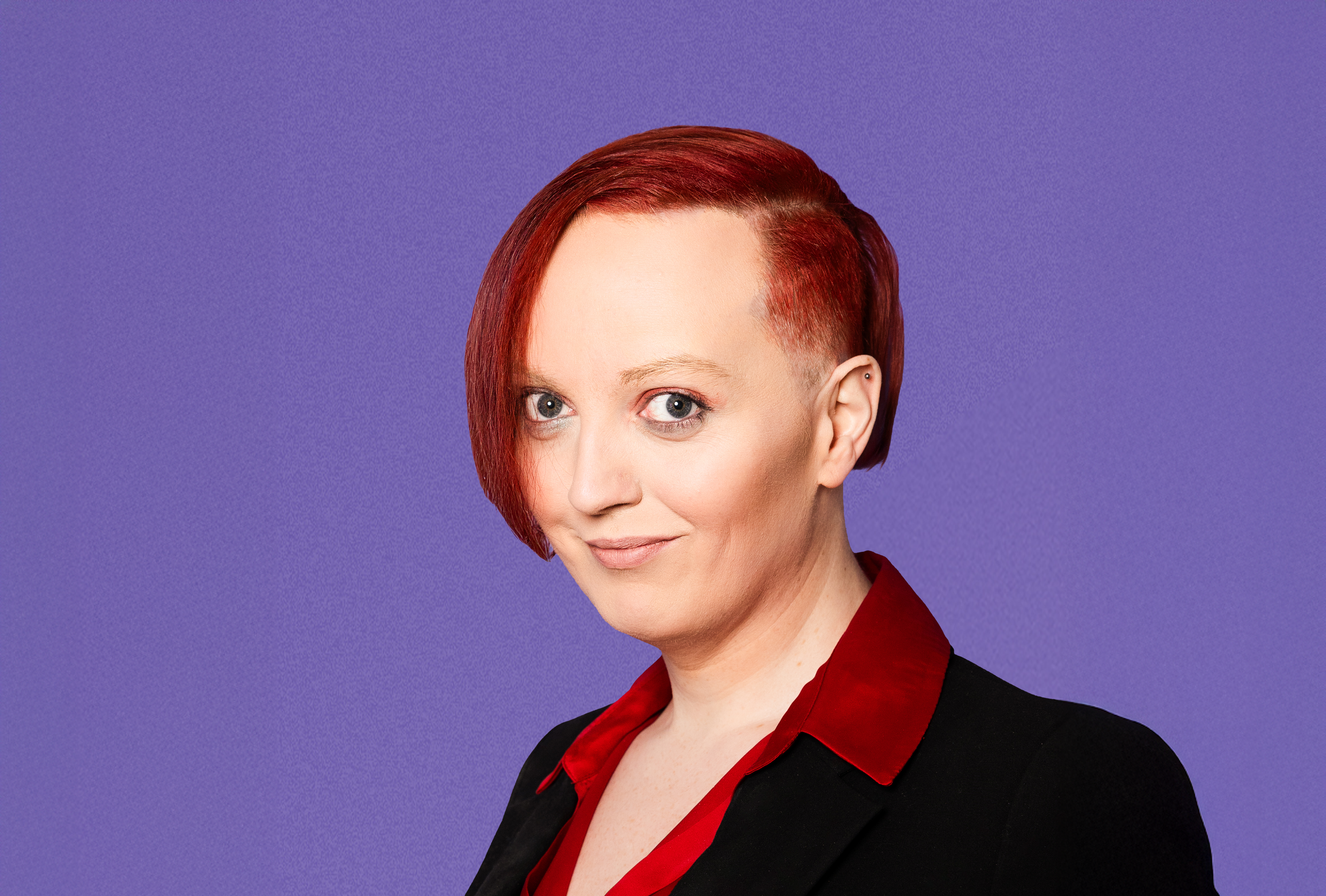 Alexandra brosandi með rautt hár stutt klippt á einni hlið, sítt á hinni í svörtum superman bol á ljósum bakgrunni.