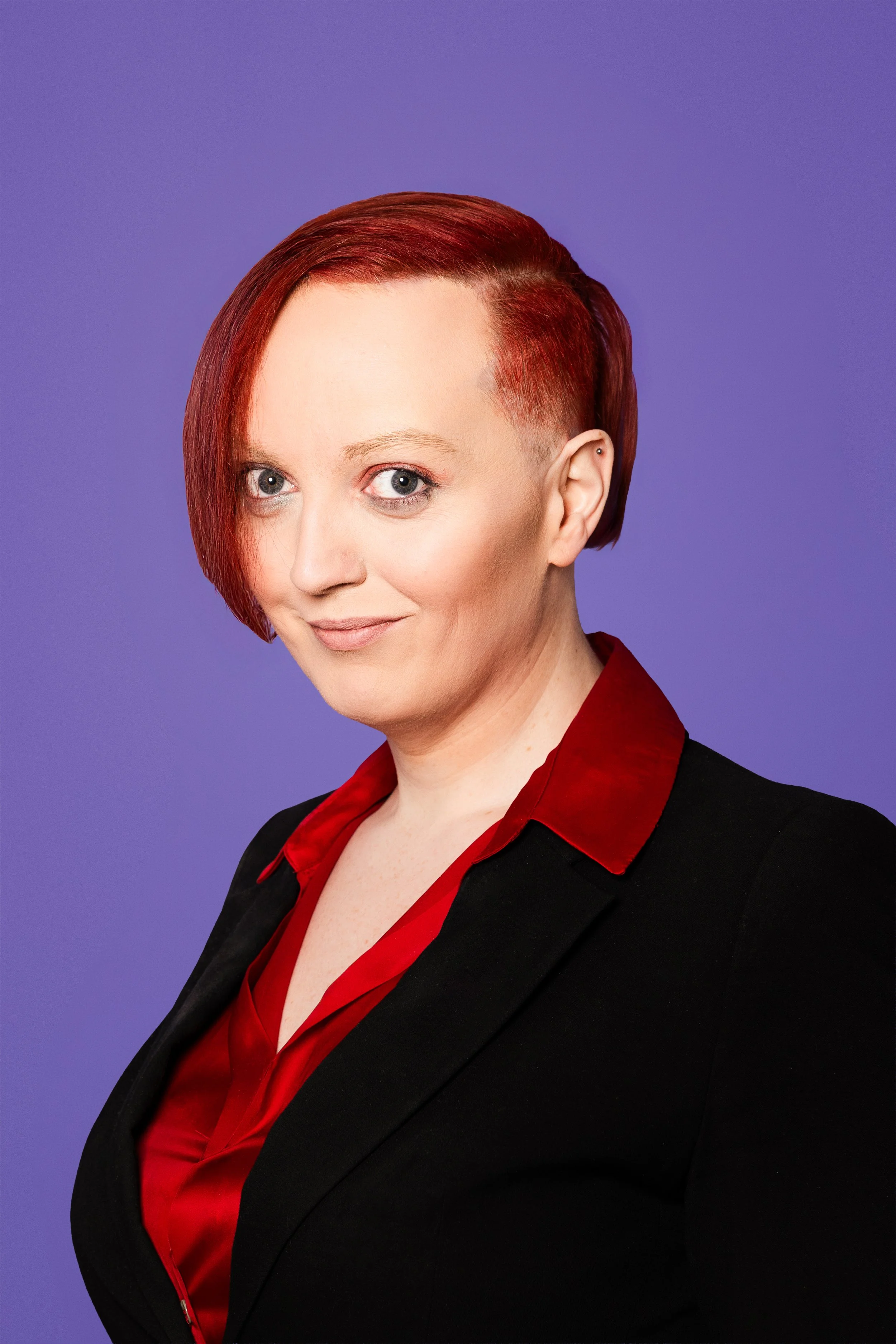 Alexandra brosandi með rautt hár stutt klippt á einni hlið, sítt á hinni í svörtum superman bol á ljósum bakgrunni.