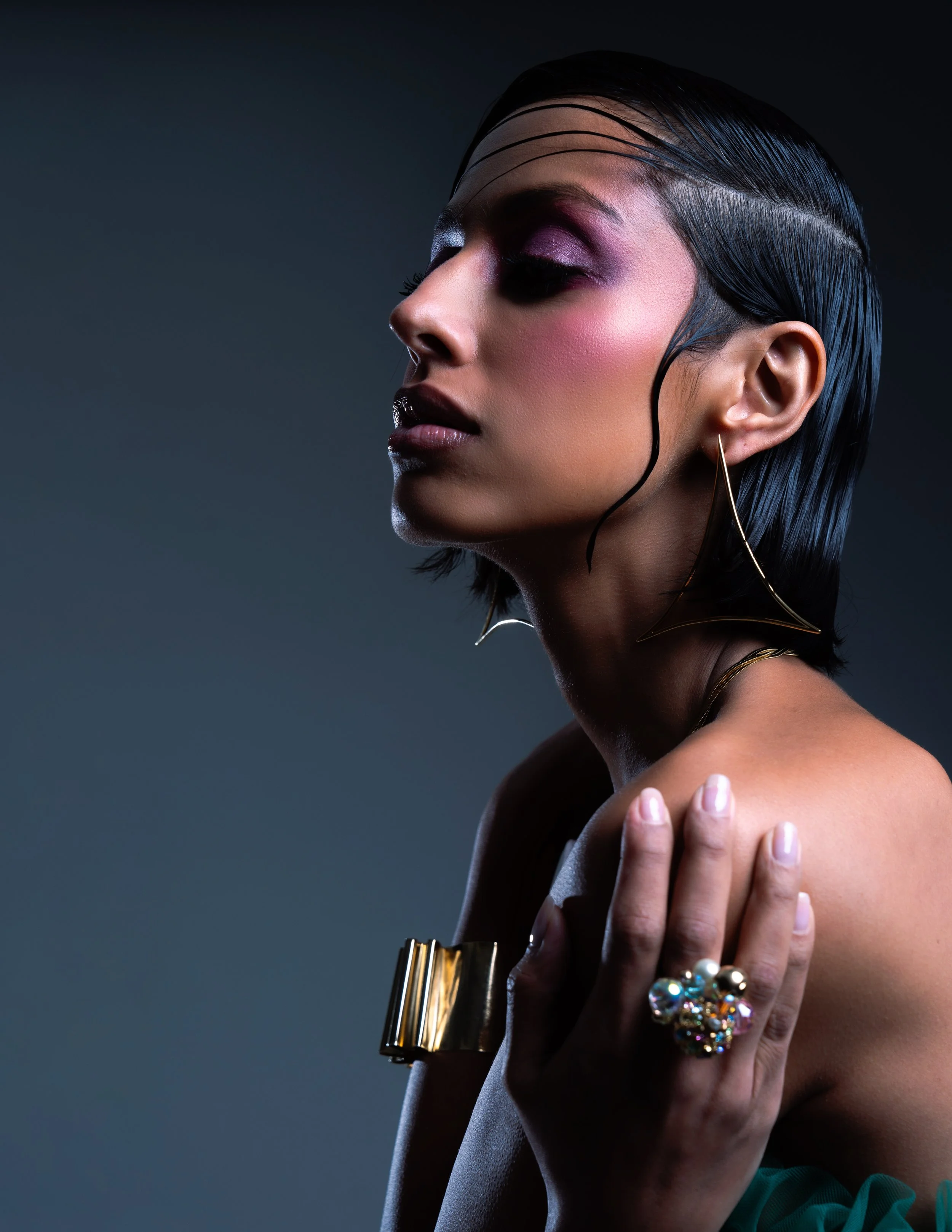 A woman with short, wet-looking black hair, elegant makeup with pink eyeshadow, and large gold jewelry, posed with her eyes closed and hand on shoulder against a dark background.