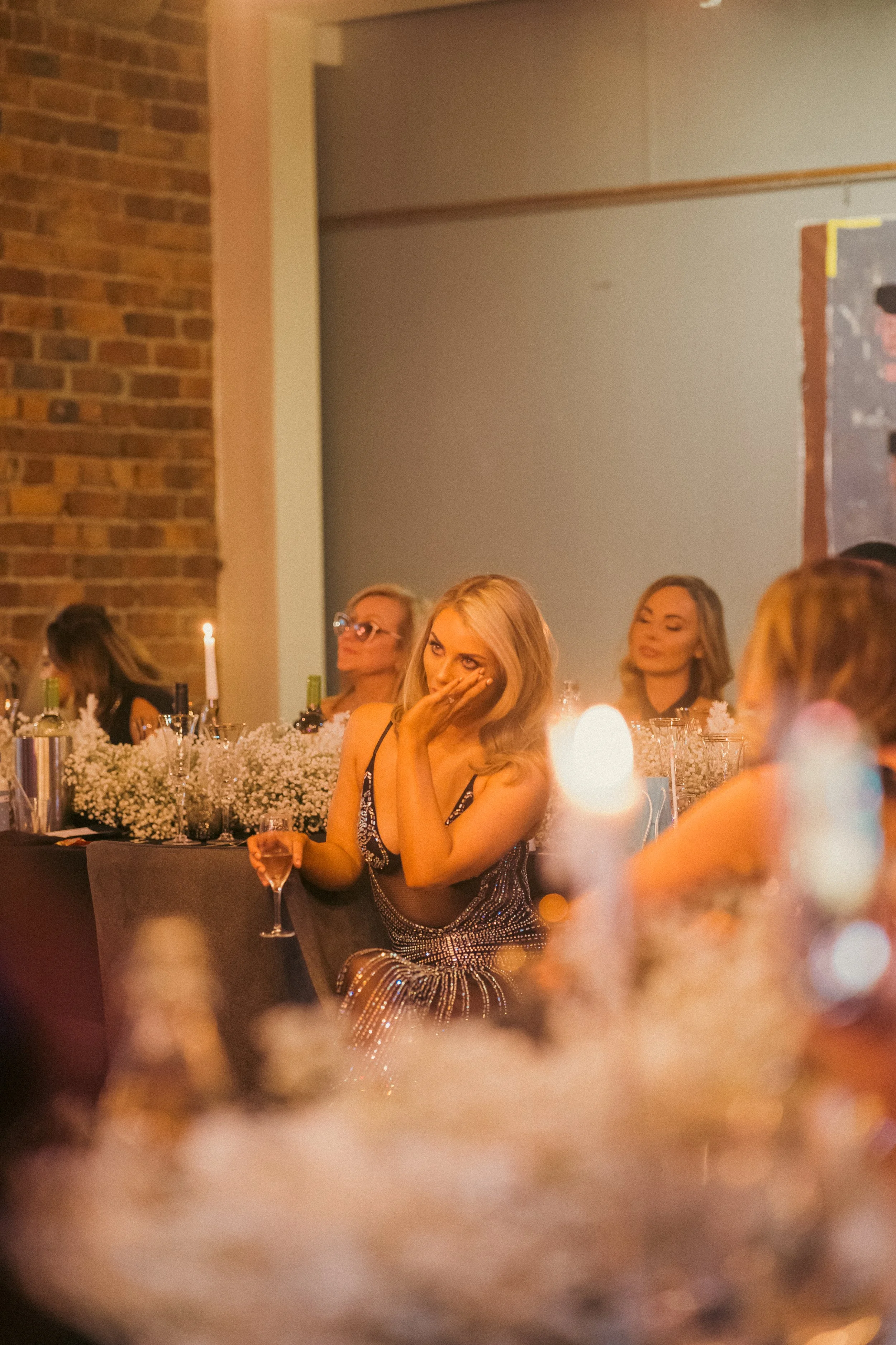 Women at a dimly lit social gathering or celebration, sitting at a decorated table with flowers, candles, and drinks, some appear emotional or thoughtful.