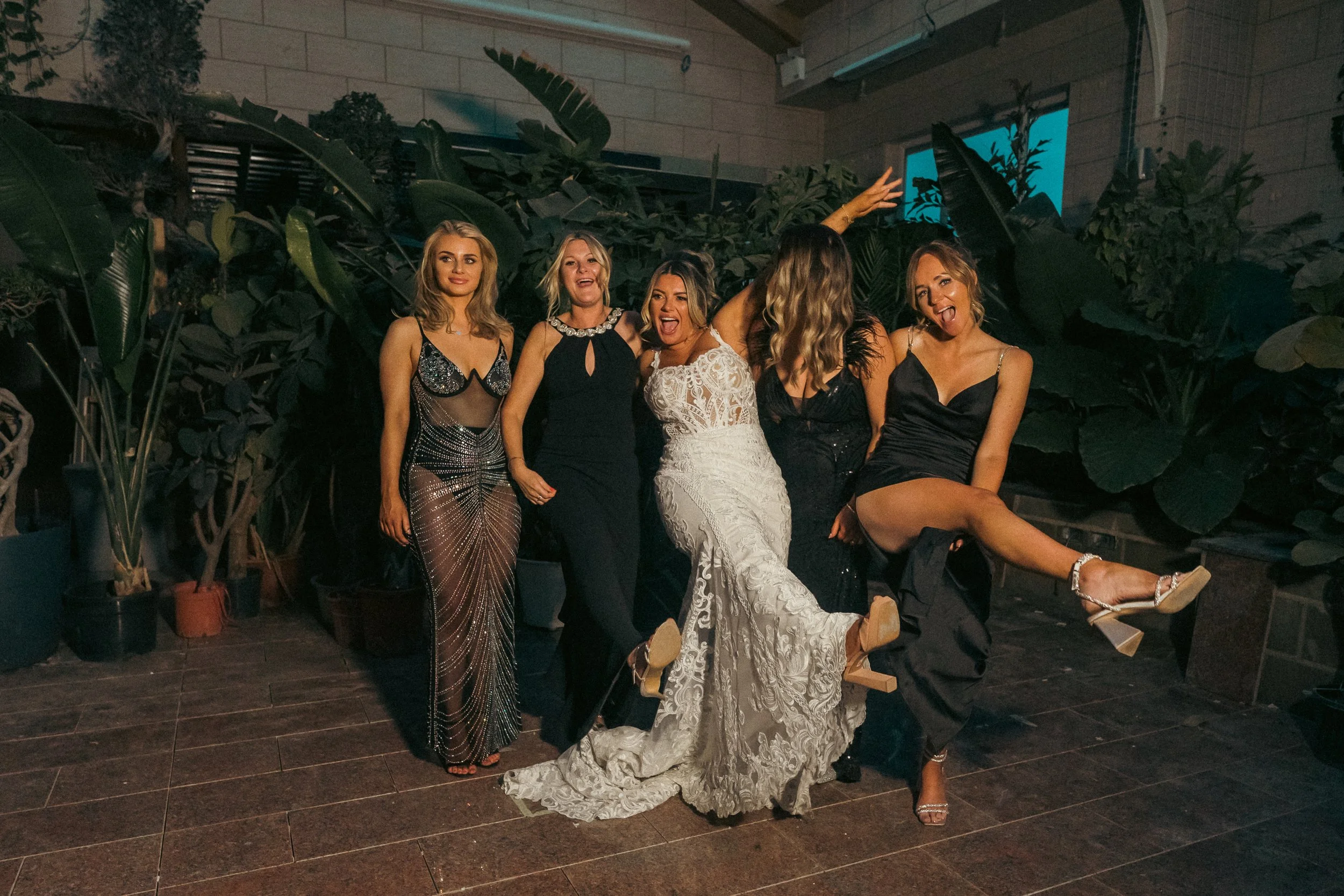 A group of six women dressed in elegant evening gowns posing together at a party or celebration, with one woman lifting her leg playfully.