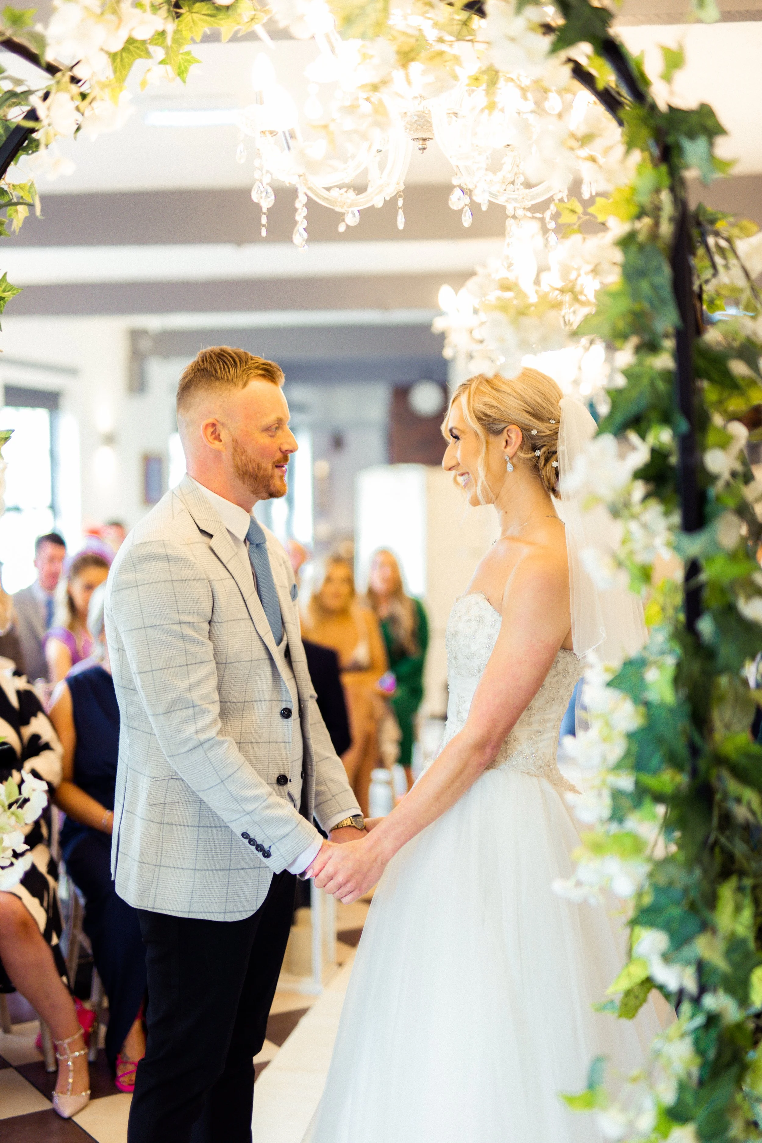 A bride and groom holding hands during their wedding ceremony, exchanging vows under a floral arch with a chandelier overhead, surrounded by guests.
