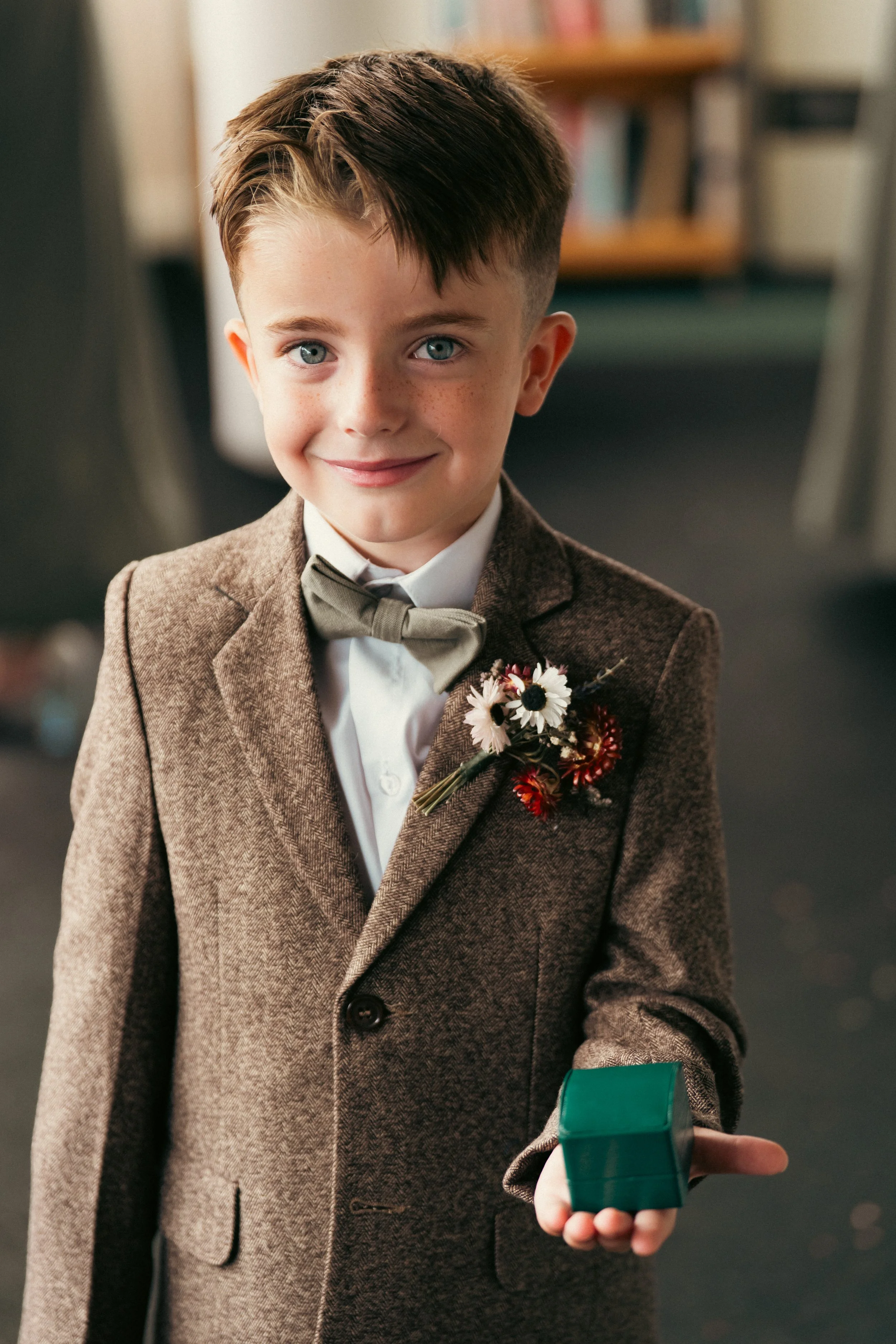 A young boy dressed in a brown suit with a bowtie, holding a small green box, smiling at the camera.