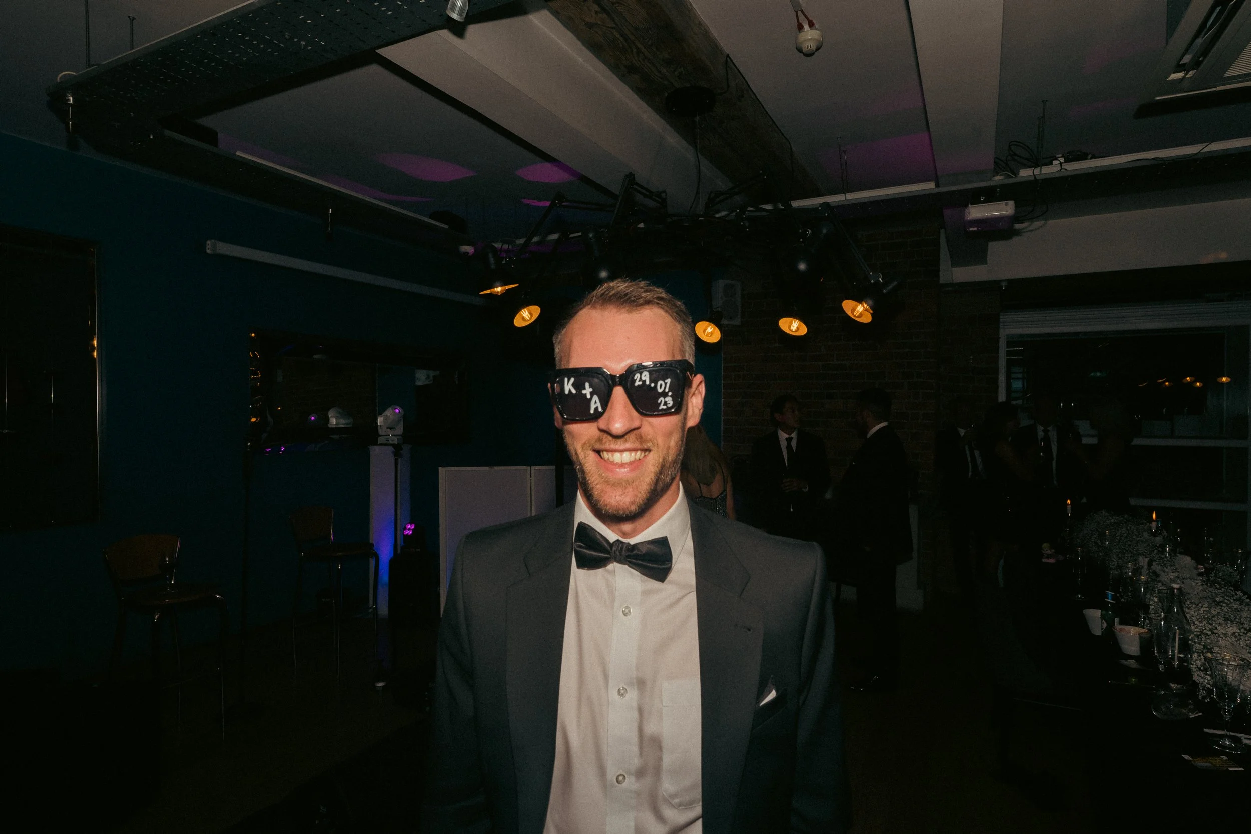 A man dressed in a tuxedo with a white shirt and black bow tie, smiling at a party. He is wearing sunglasses with writing on the lenses. The background shows a dimly lit indoor venue with overhead lights, a table with drinks, and other guests in form