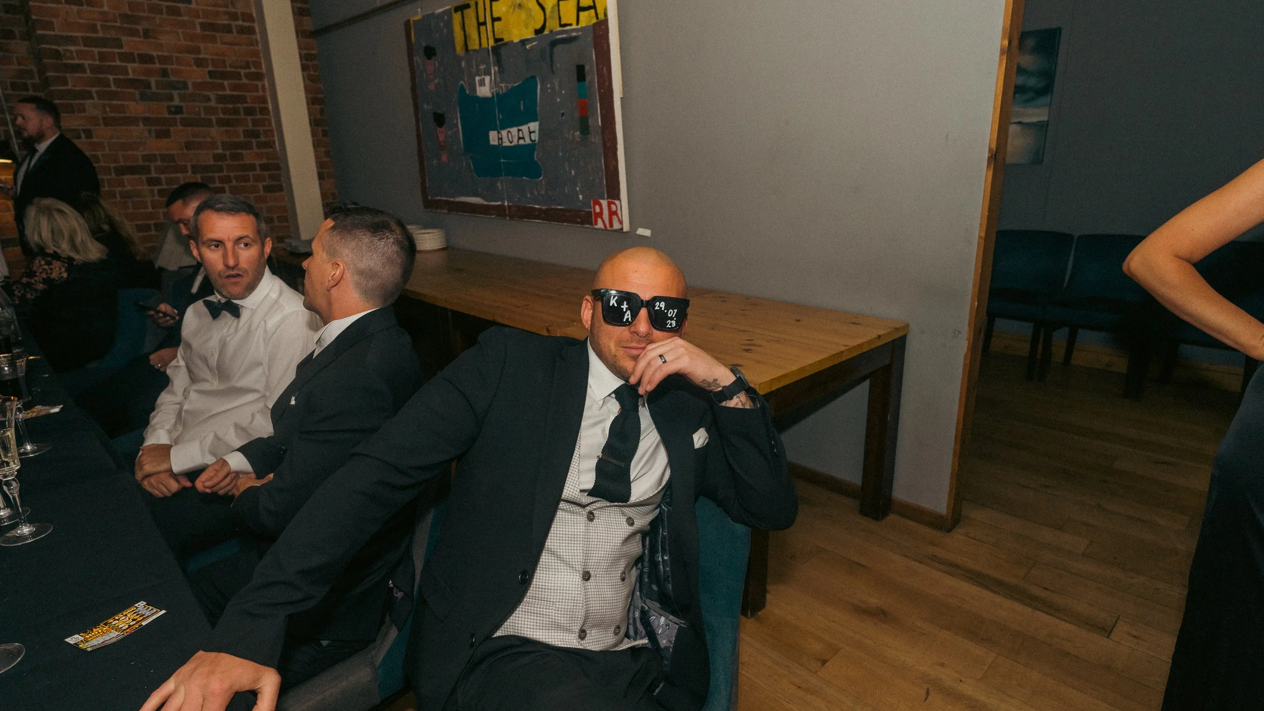 Man wearing custom black sunglasses, black suit, white shirt, black tie, and vest, sitting at a dinner event with others, in a room with brick wall and abstract artwork.