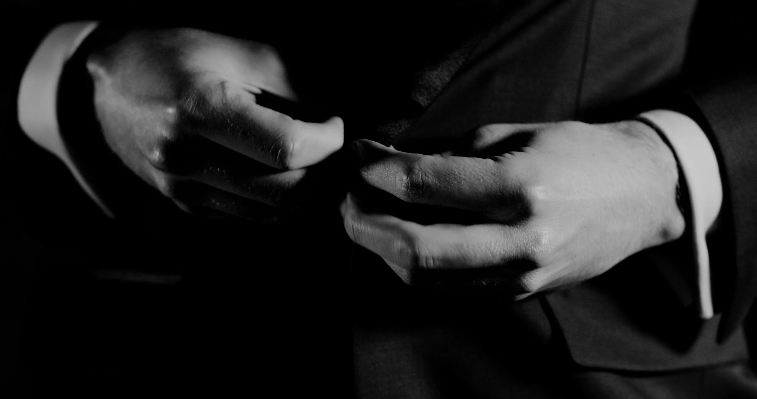 Close-up black and white photo of a person adjusting their suit jacket, focusing on hands and part of the suit.