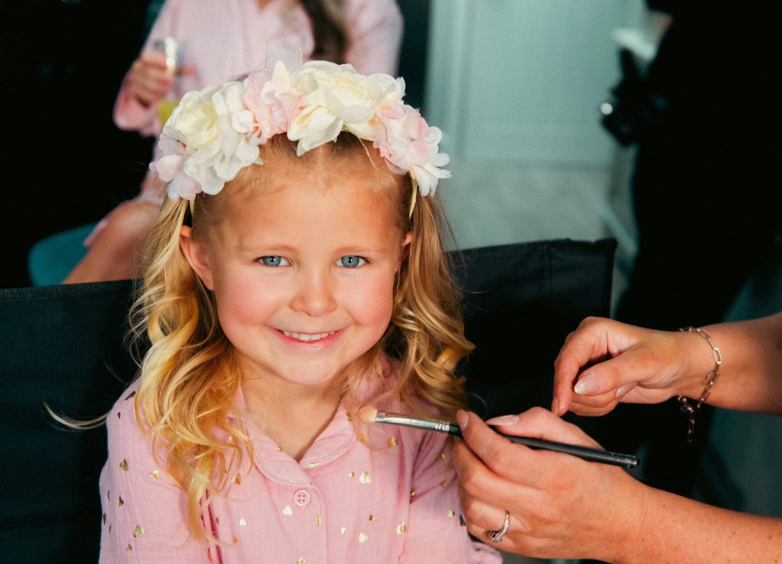 A young girl with blonde hair and blue eyes, wearing a pink shirt with gold hearts and a floral headband, is smiling while getting her face painted by an artist. The artist's hands are visible, holding a paintbrush near her face. Other people are in 