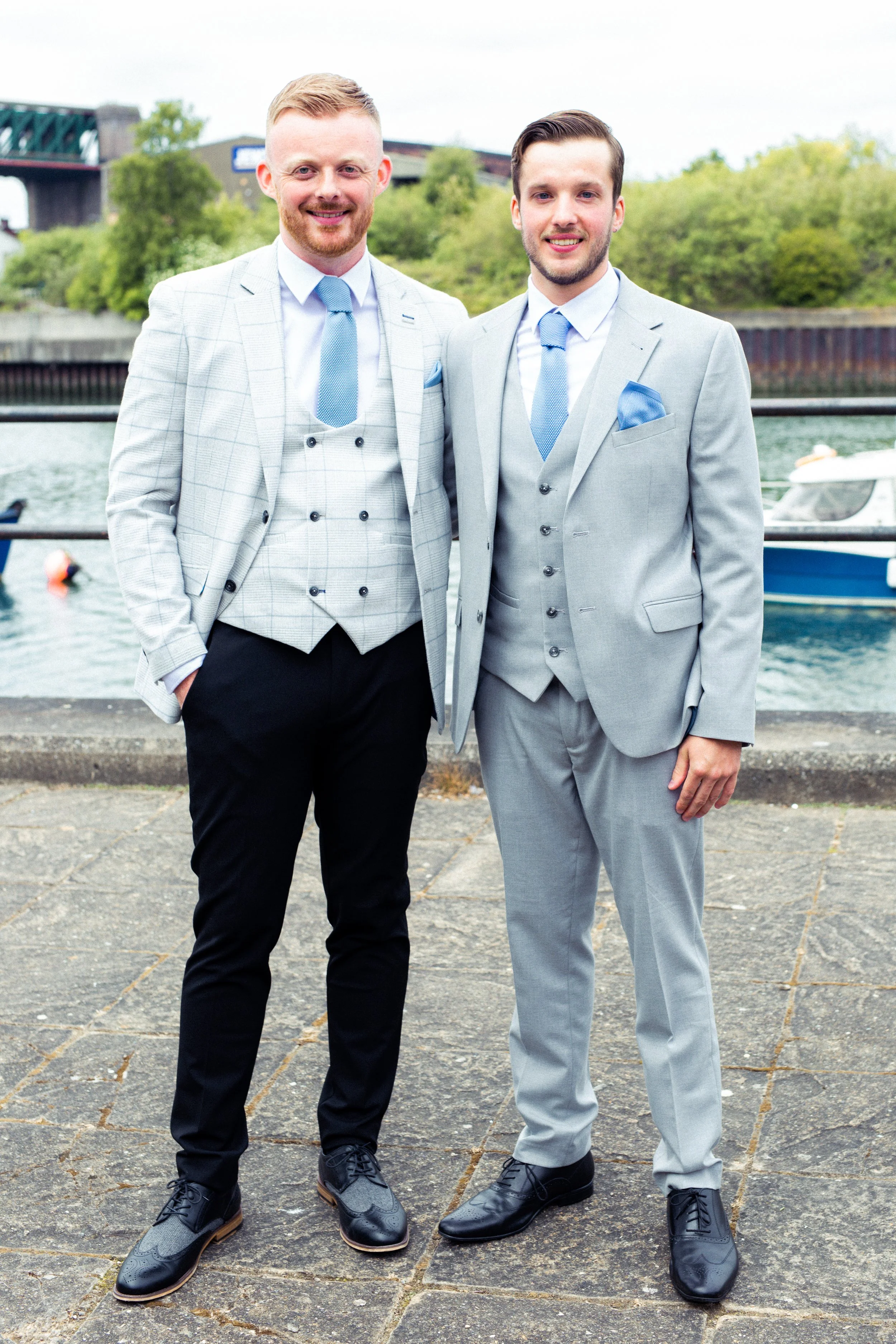 Two men in suits standing outdoors near water with boats and bridge in the background.