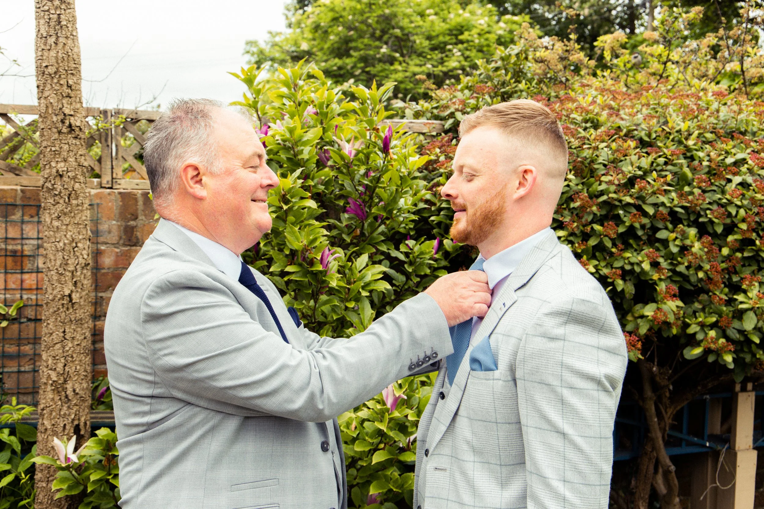 An older man helping a younger man adjust his tie outdoors, both dressed in light gray suits, with greenery and flowering bushes in the background.