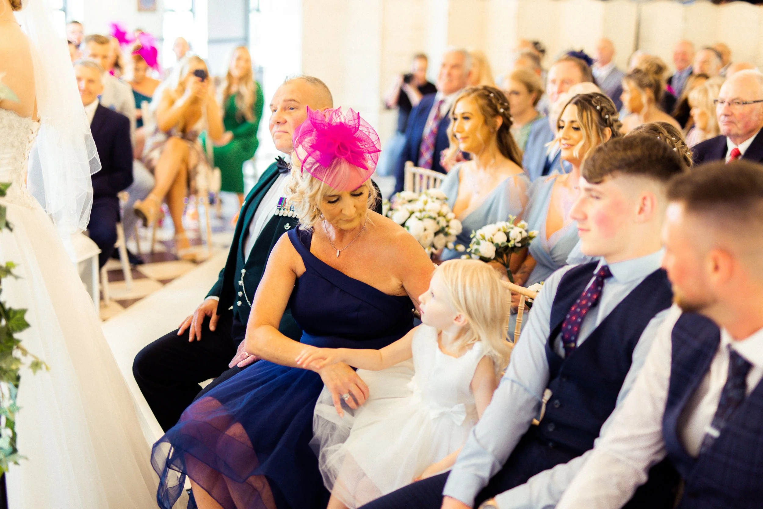 Wedding ceremony with family and guests seated, a woman in a navy dress and pink fascinator looks at a young girl in a white dress, sitting next to a man in a dark suit and tie, with bridesmaids holding bouquets in the background.