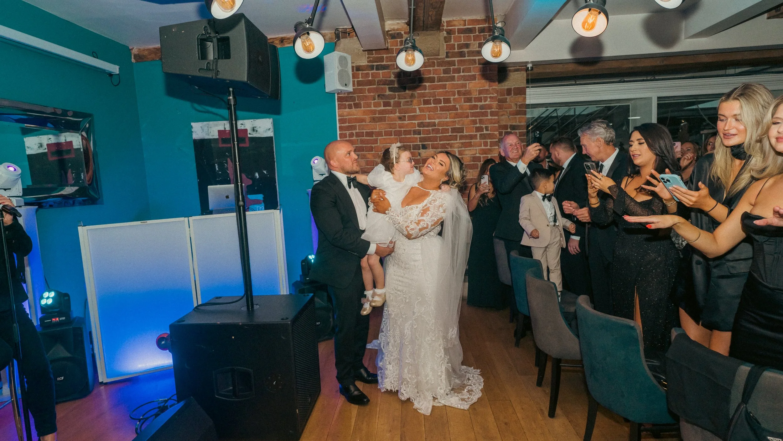 Wedding reception with a bride and groom holding a young girl, surrounded by guests taking photos in a decorated indoor venue.