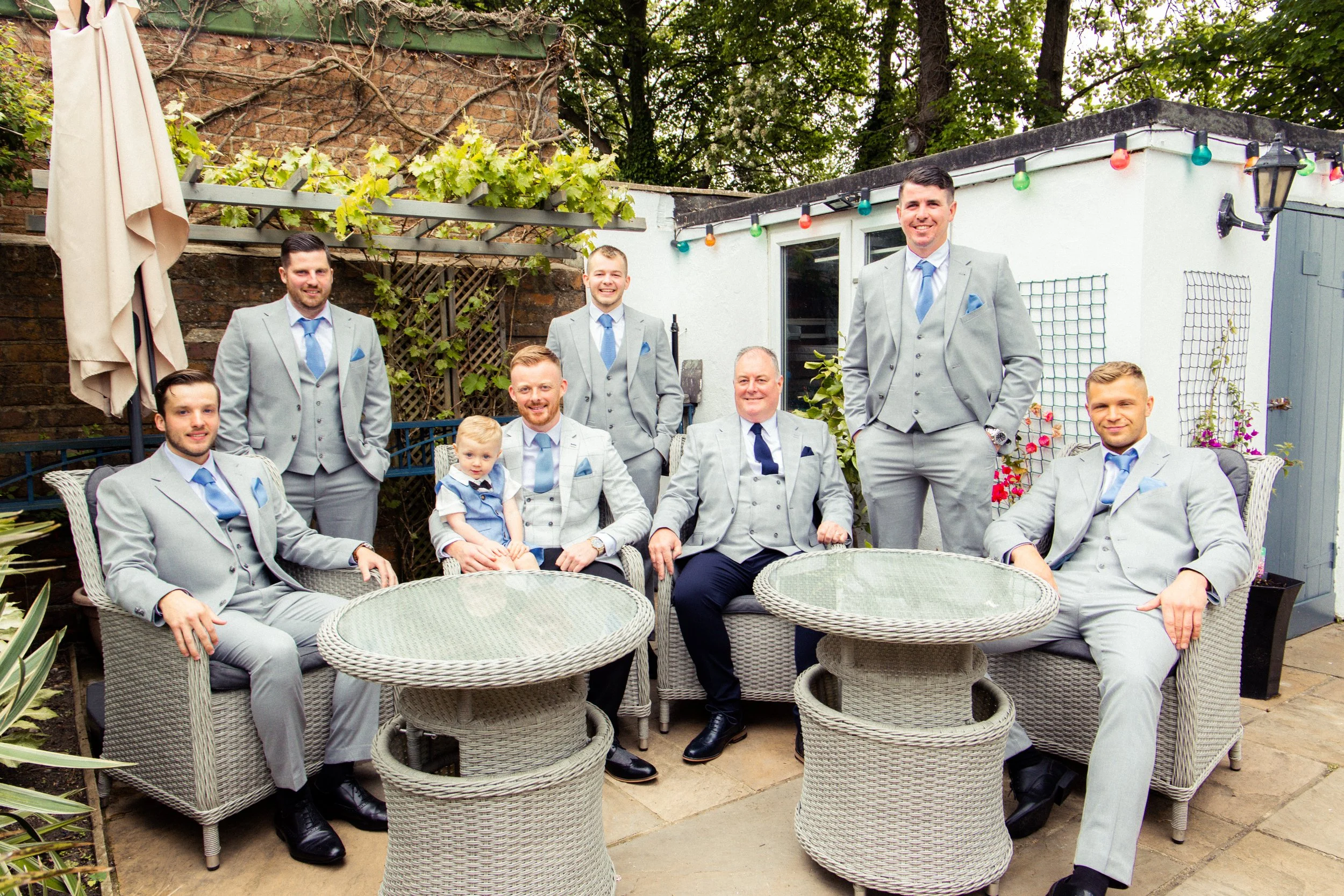 A group of nine men and one young boy dressed in light gray suits with blue ties, sitting and standing in a backyard patio with outdoor furniture and greenery.