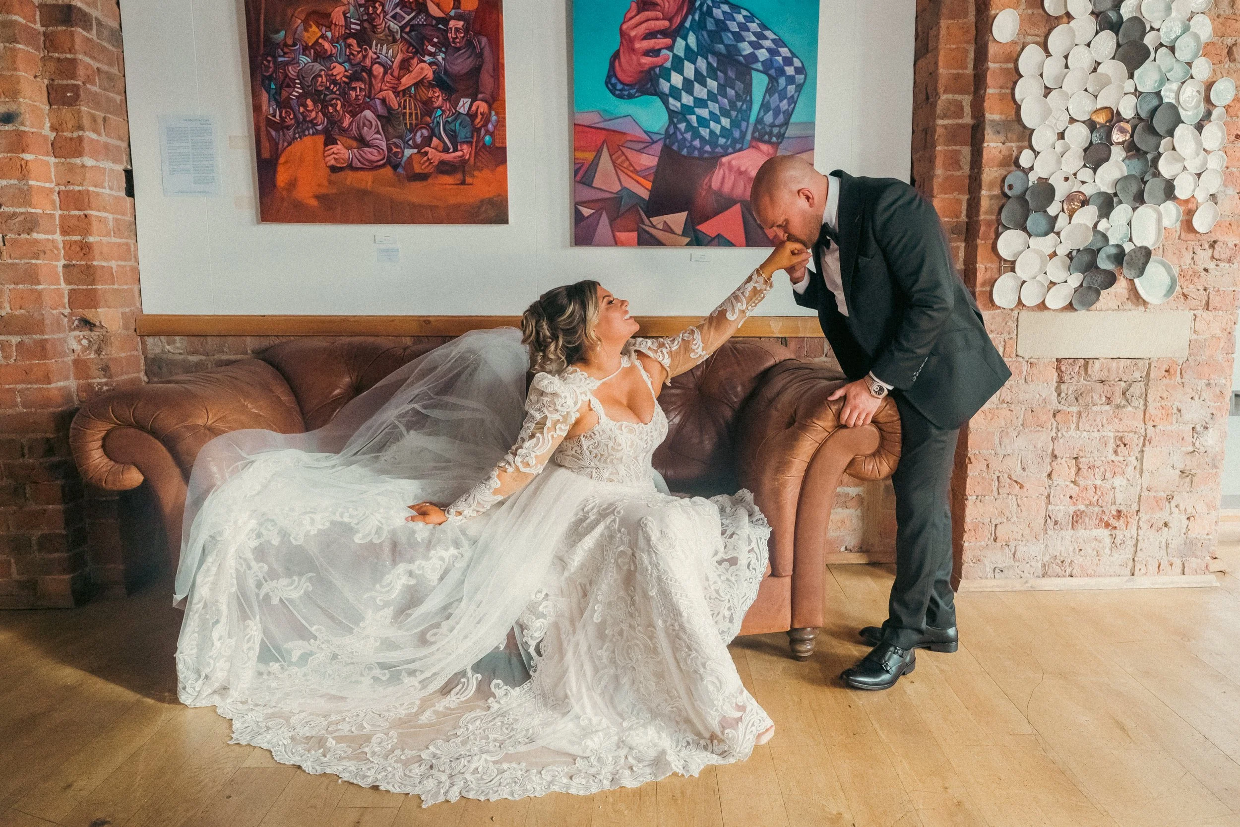 A bride in a white lace wedding dress sitting on a brown leather couch, reaching out to kiss a groom in a black tuxedo who is leaning over the armrest, kissing her hand, in an art gallery with brick walls and colorful paintings.