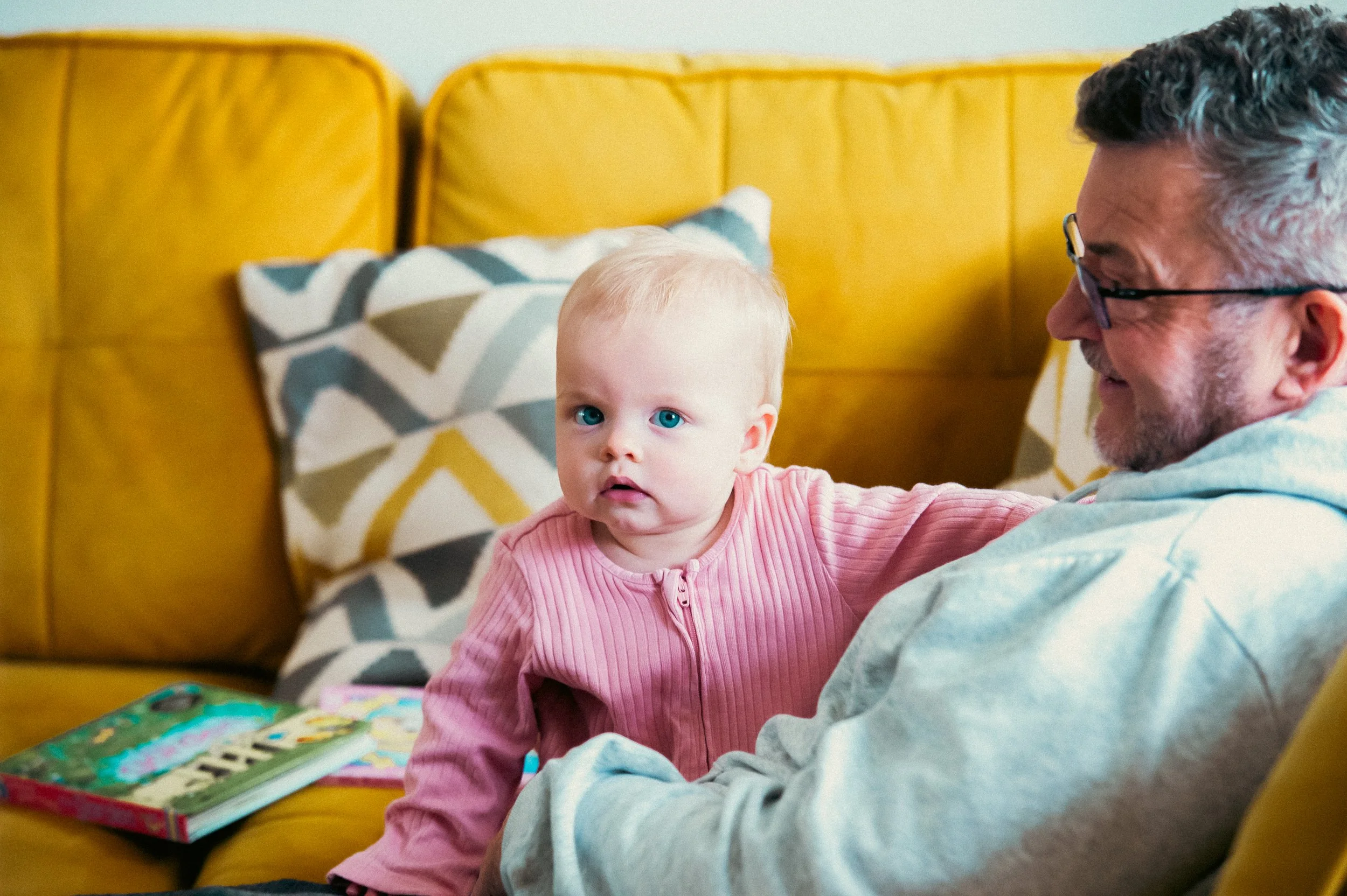A young child in a pink zip-up sweater sitting on a yellow couch next to an older man with glasses, both smiling, with colorful pillows behind them.