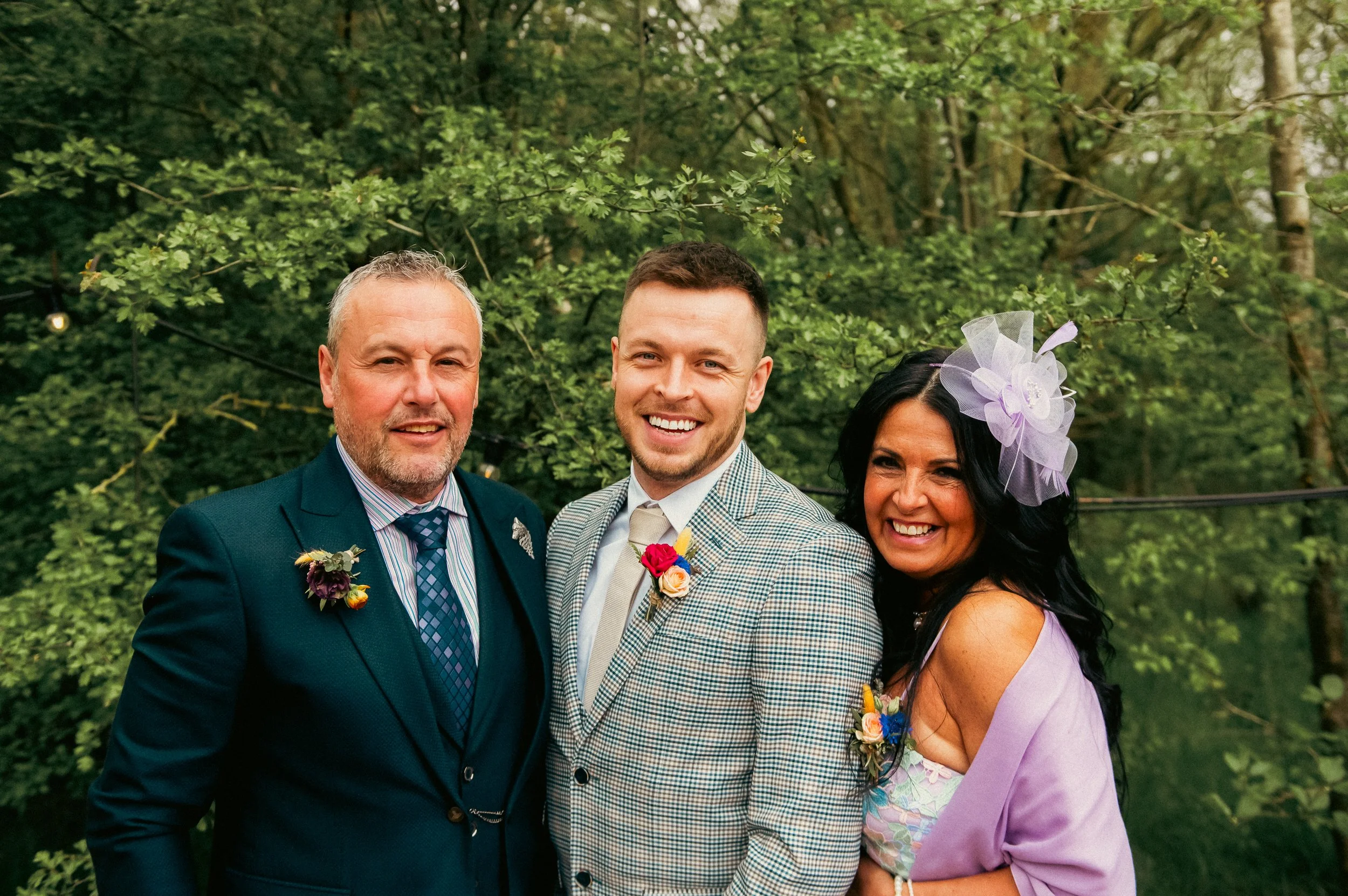 Three people at an outdoor wedding, all dressed in formal attire, smiling and posing for a photo in a green, wooded setting, with flowers and decorative elements on their outfits.