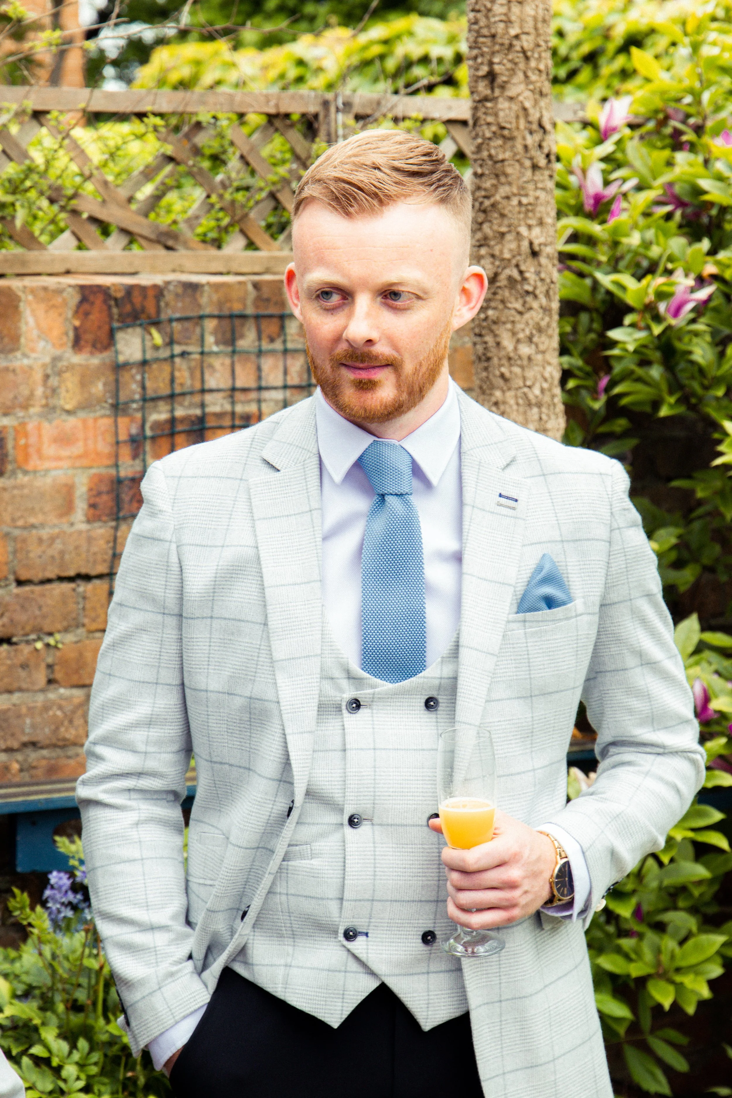 A man with red hair and a beard wearing a light gray plaid suit, blue tie, and white shirt, holding a glass of orange drink, standing outdoors with a brick wall, wooden fence, green foliage, and purple flowers in the background.