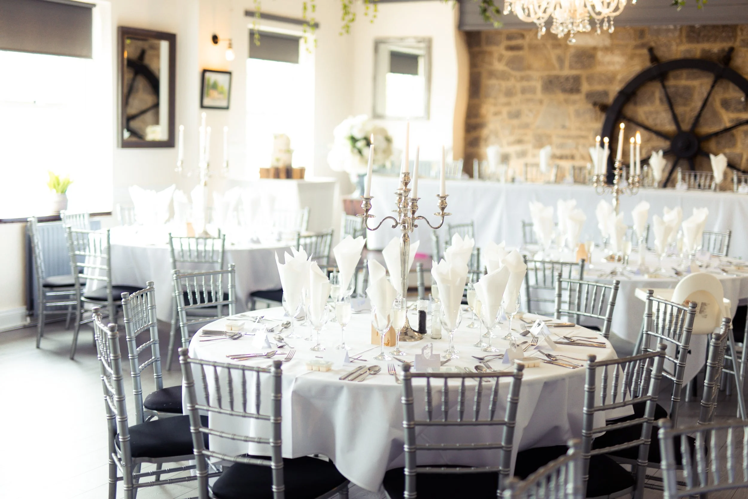 An elegantly set round table decorated for a formal event in a bright room with large windows, silver chairs, white tablecloths, folded napkins, silverware, wine glasses, and silver candelabras.