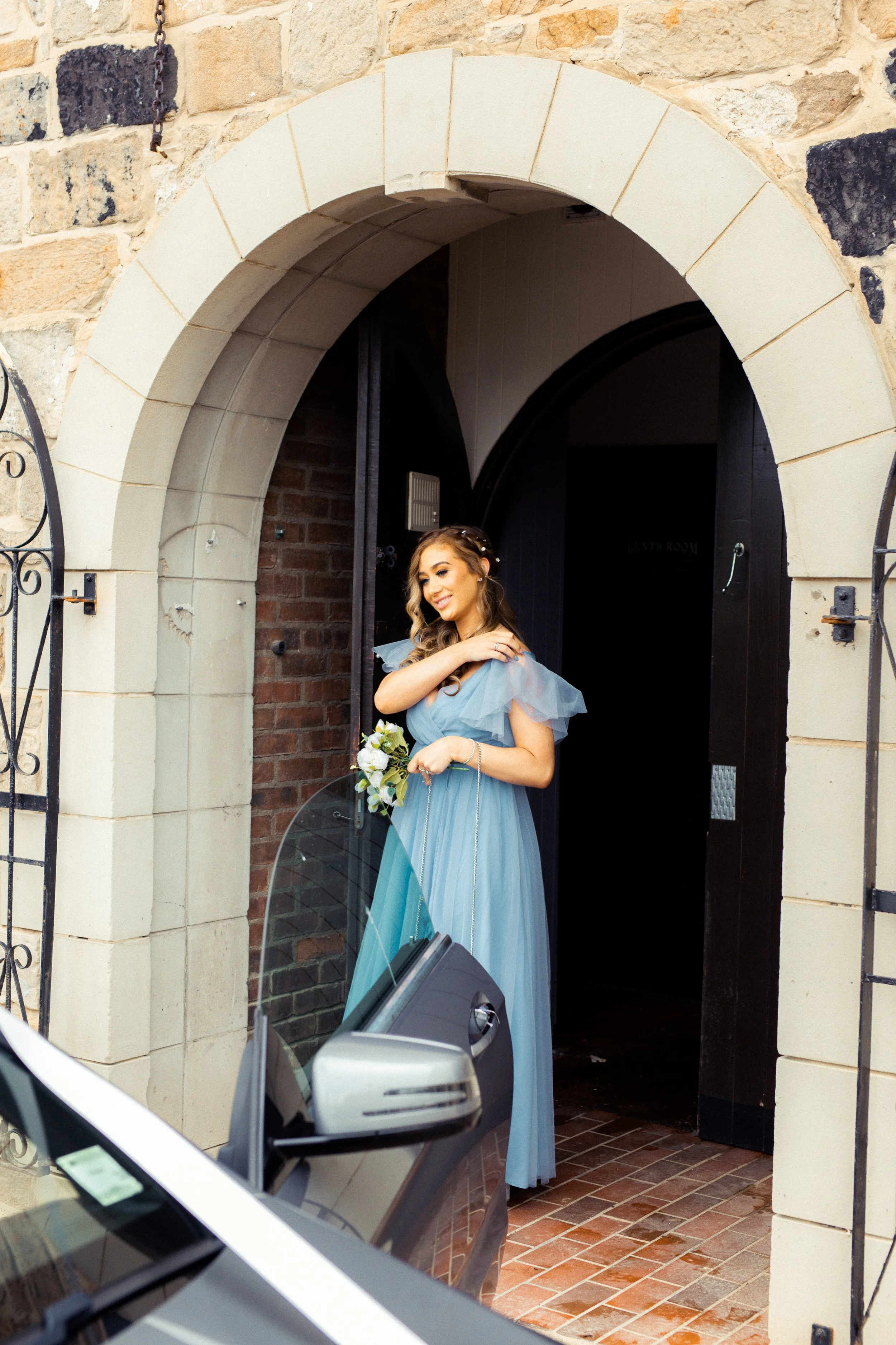 A woman dressed in a blue gown holding a bouquet of flowers standing at an arched doorway.