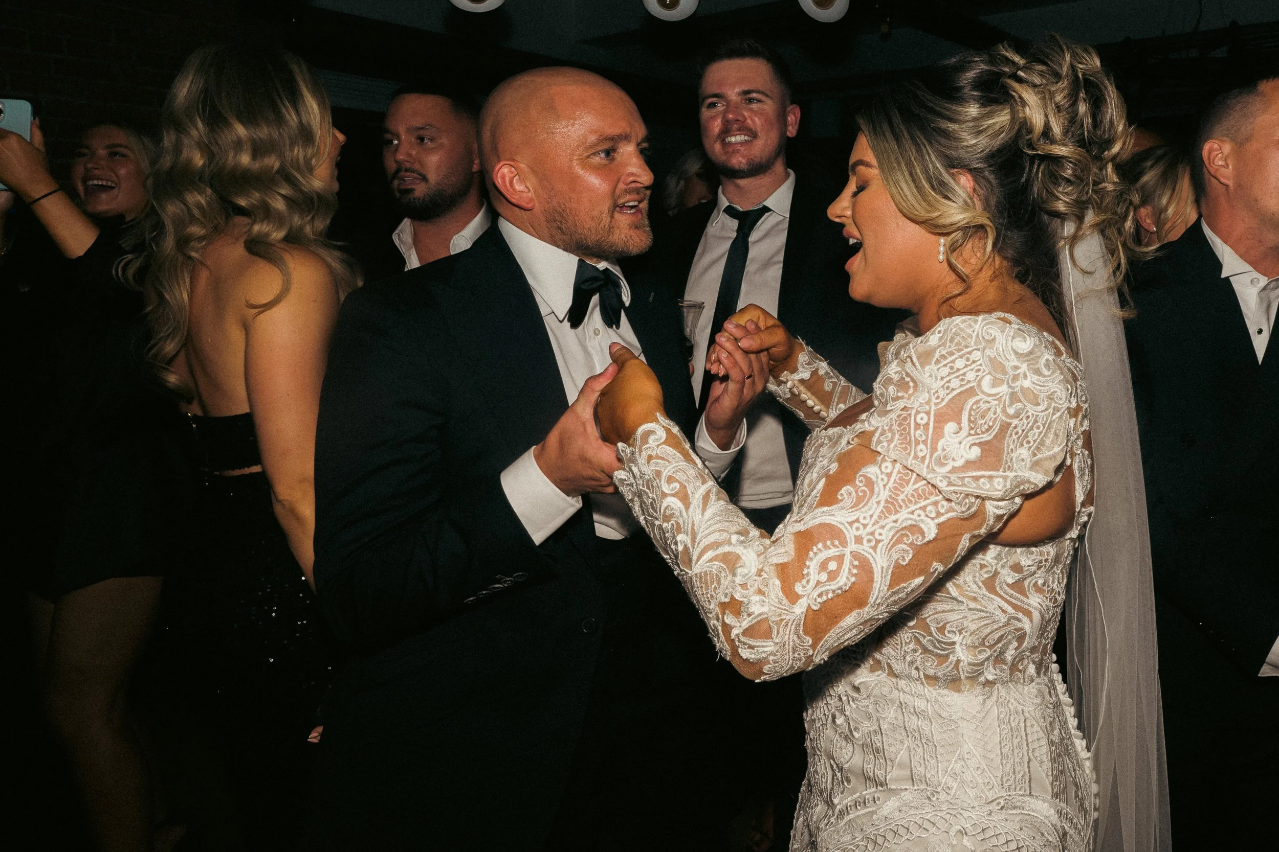 A bride and groom share a dance at their wedding reception, holding hands and smiling, surrounded by guests in formal attire.