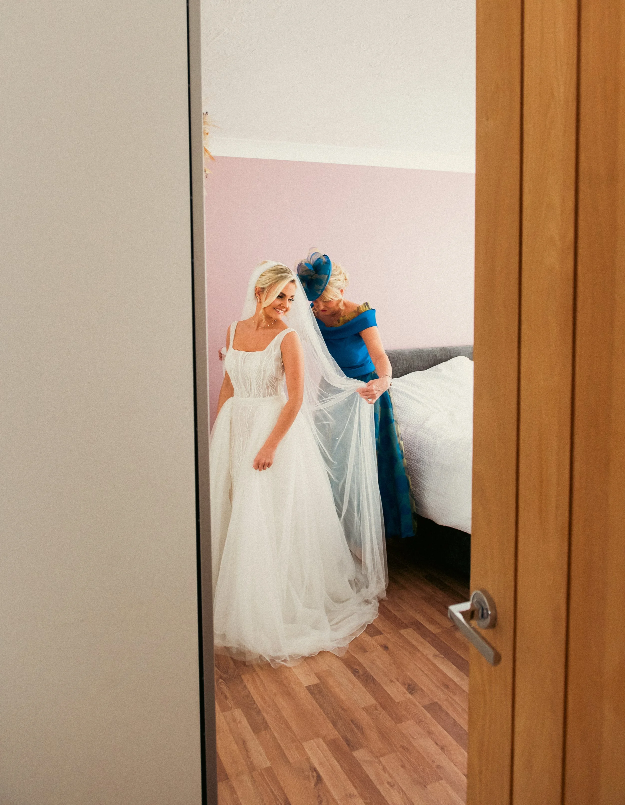 A bride in a white wedding dress and veil smiling as an older woman in a blue dress helps her with her gown in a bedroom.