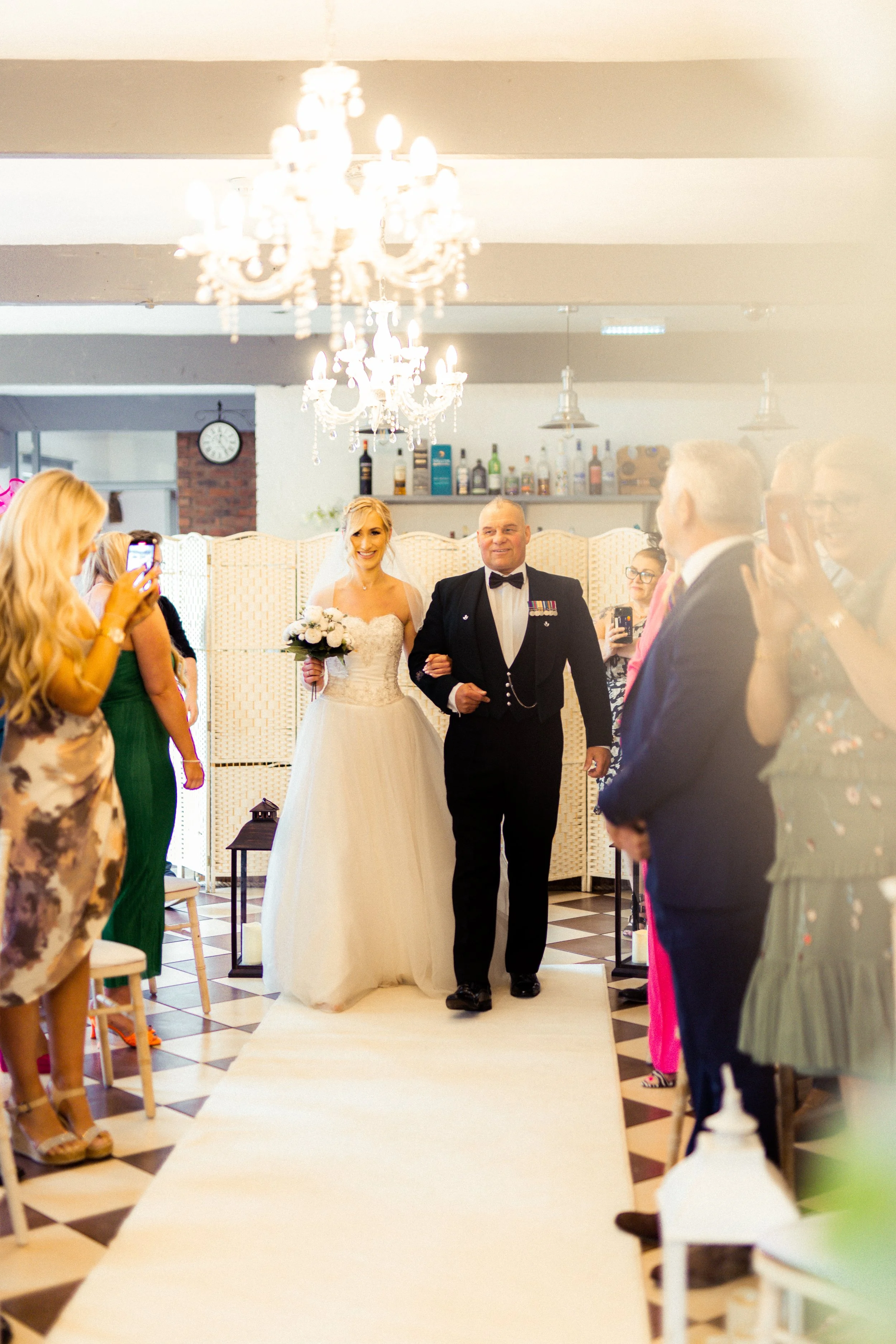 A bride walking down the aisle with a man, likely her father, at a wedding reception with guests taking photos.
