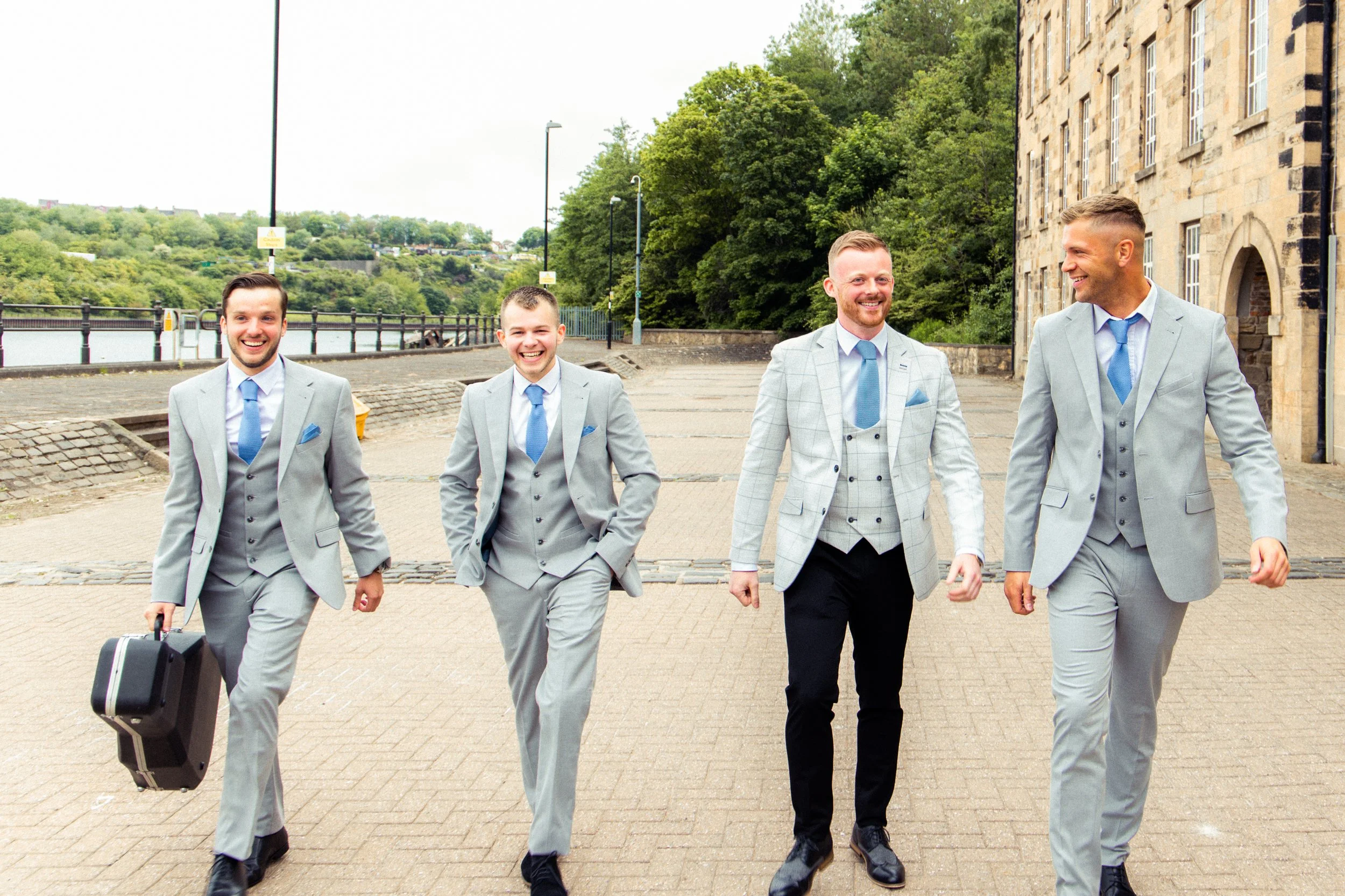 Four men in suits walking outdoors on a paved pathway, smiling and carrying briefcases, with a river, trees, and historic buildings in the background.