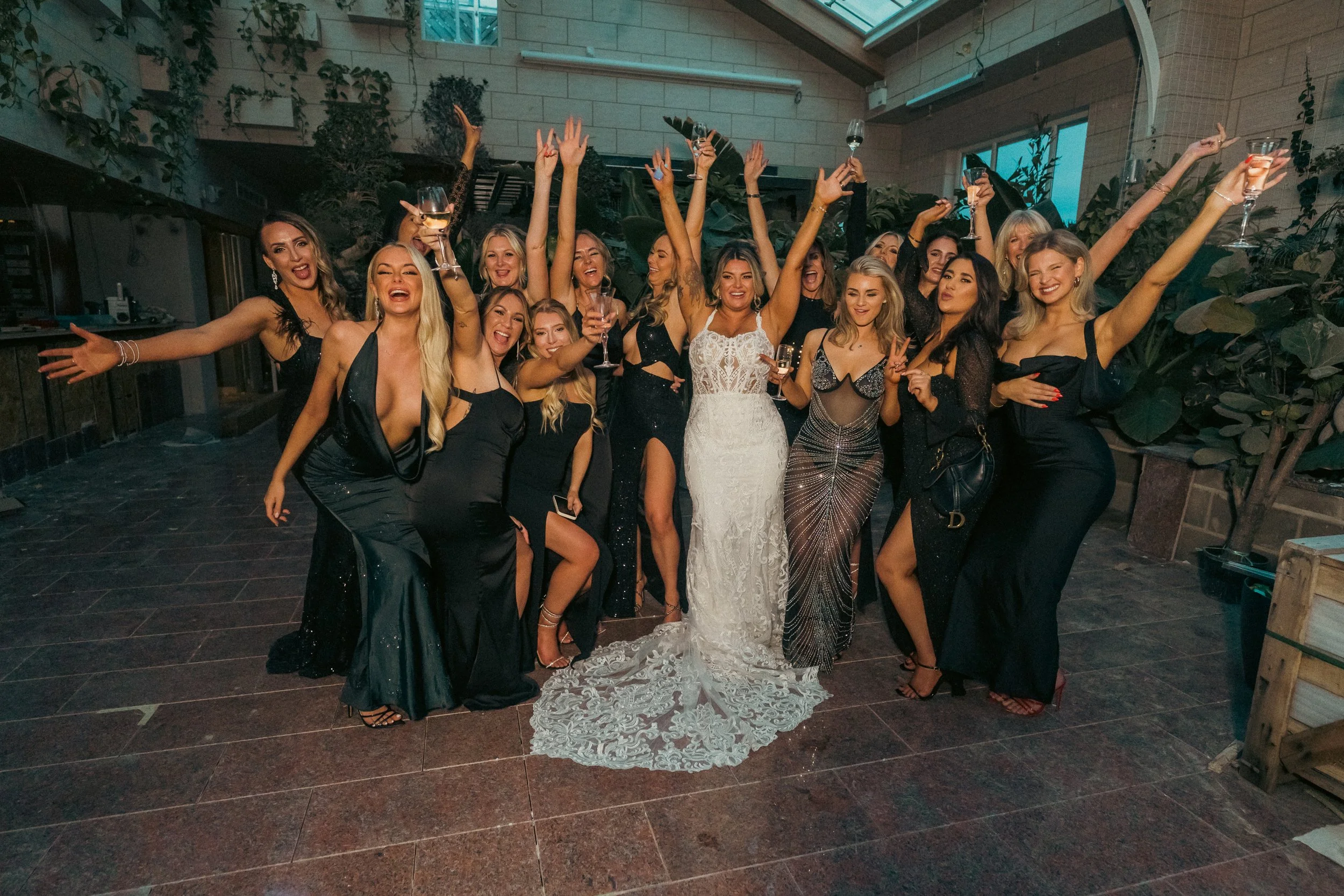 Group of women celebrating at a wedding reception inside a venue with plants and large windows, many holding glasses of wine and smiling.