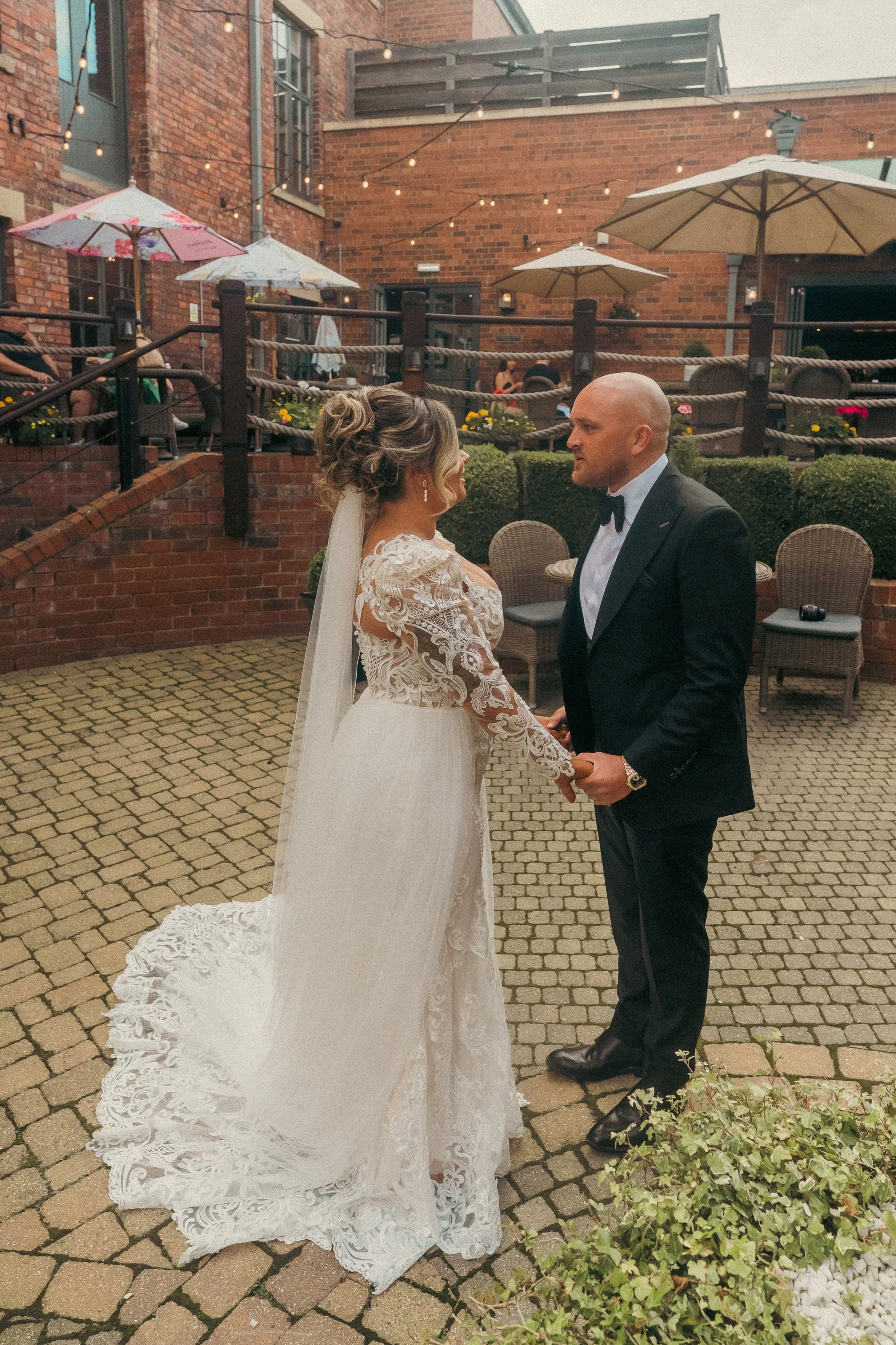 A bride and groom holding hands during their wedding ceremony outdoors, with brick buildings, string lights, umbrellas, and guests in the background.