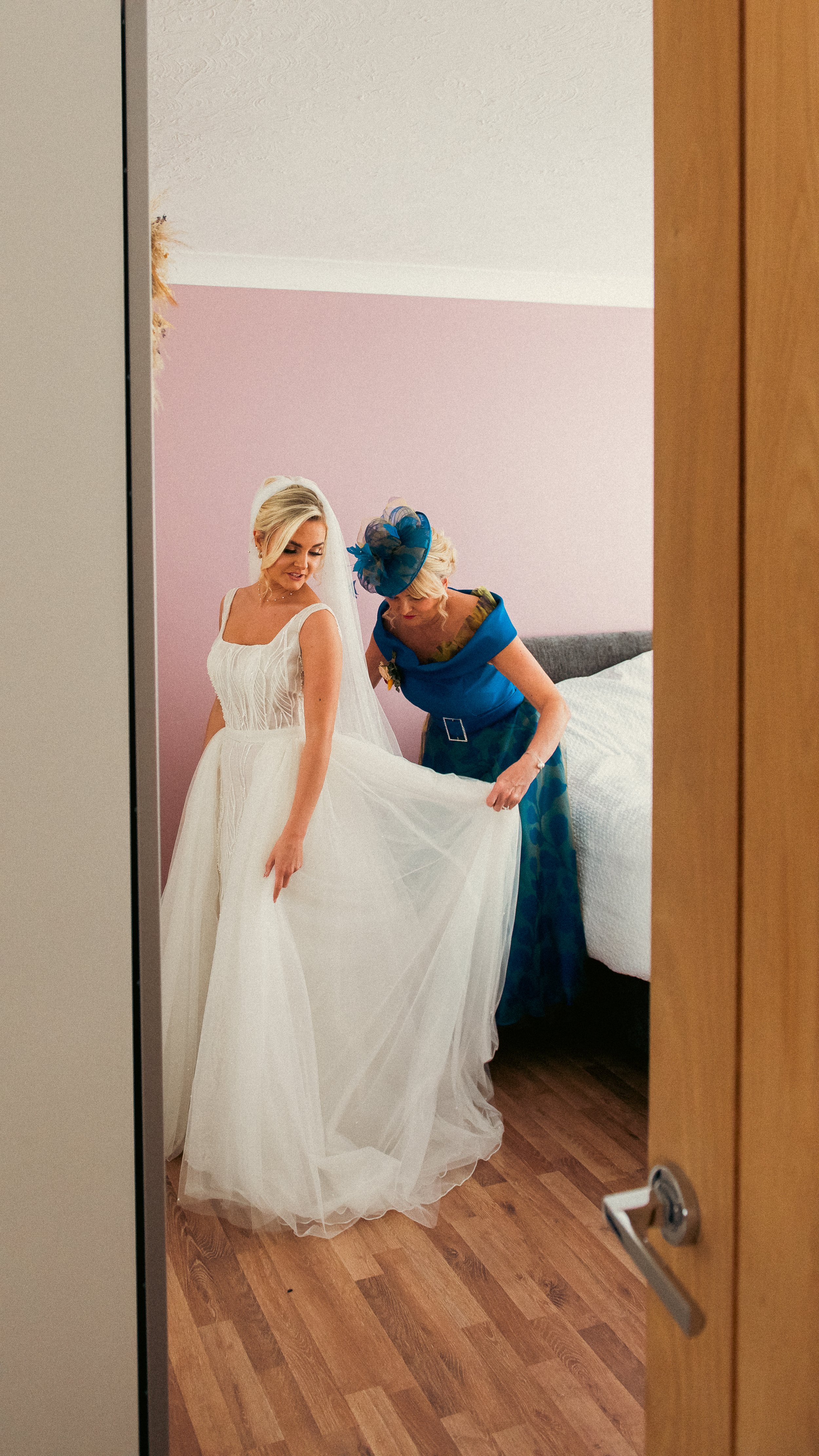 A bride in a white wedding dress getting ready with help from an older woman in a blue dress and blue hat in a bedroom with pink walls.