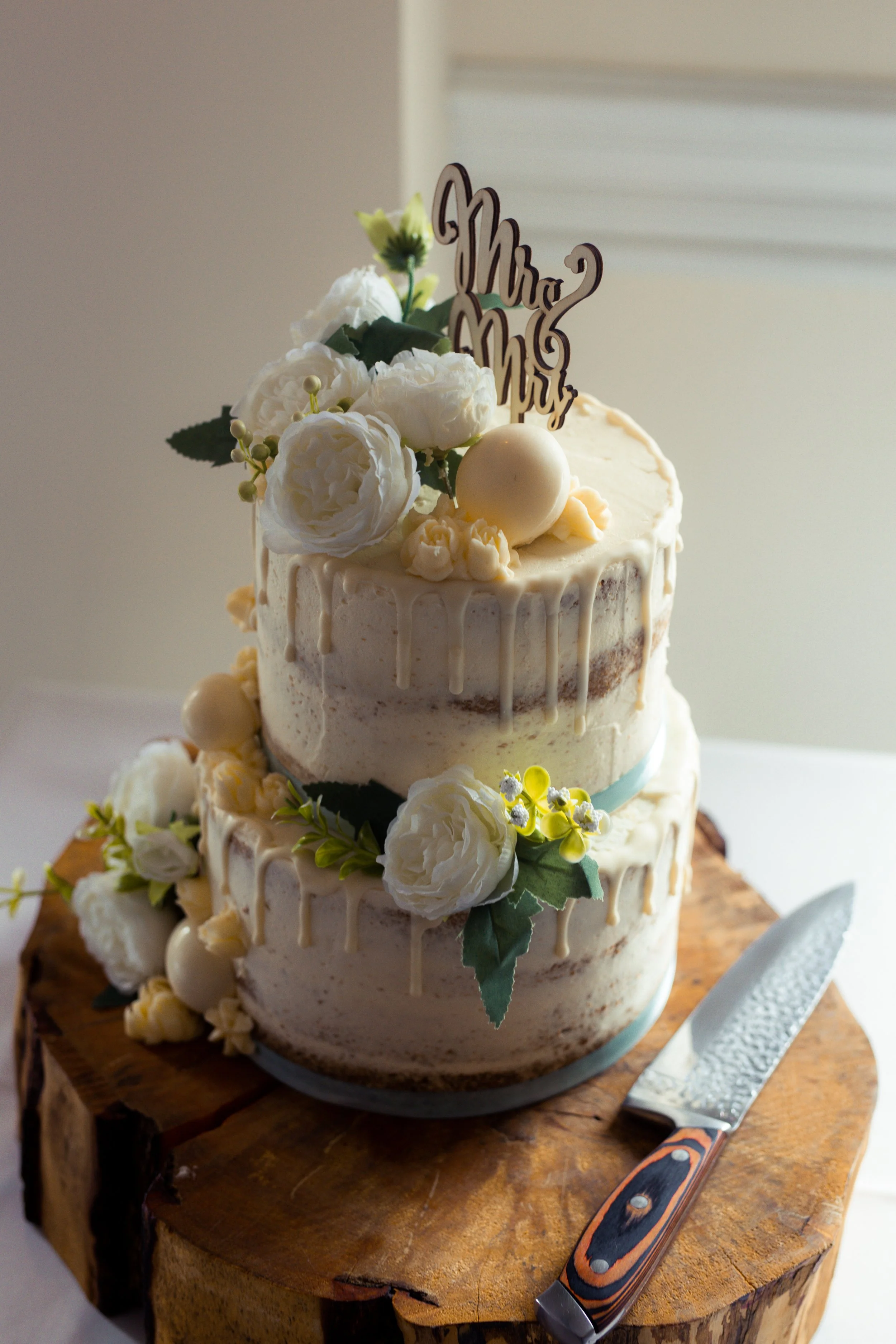 A two-tier wedding cake with white and cream-colored frosting, decorated with white flowers, green leaves, and a topper that reads 'Mrs. Mrs.' Sitting on a wooden slab with a knife beside it.