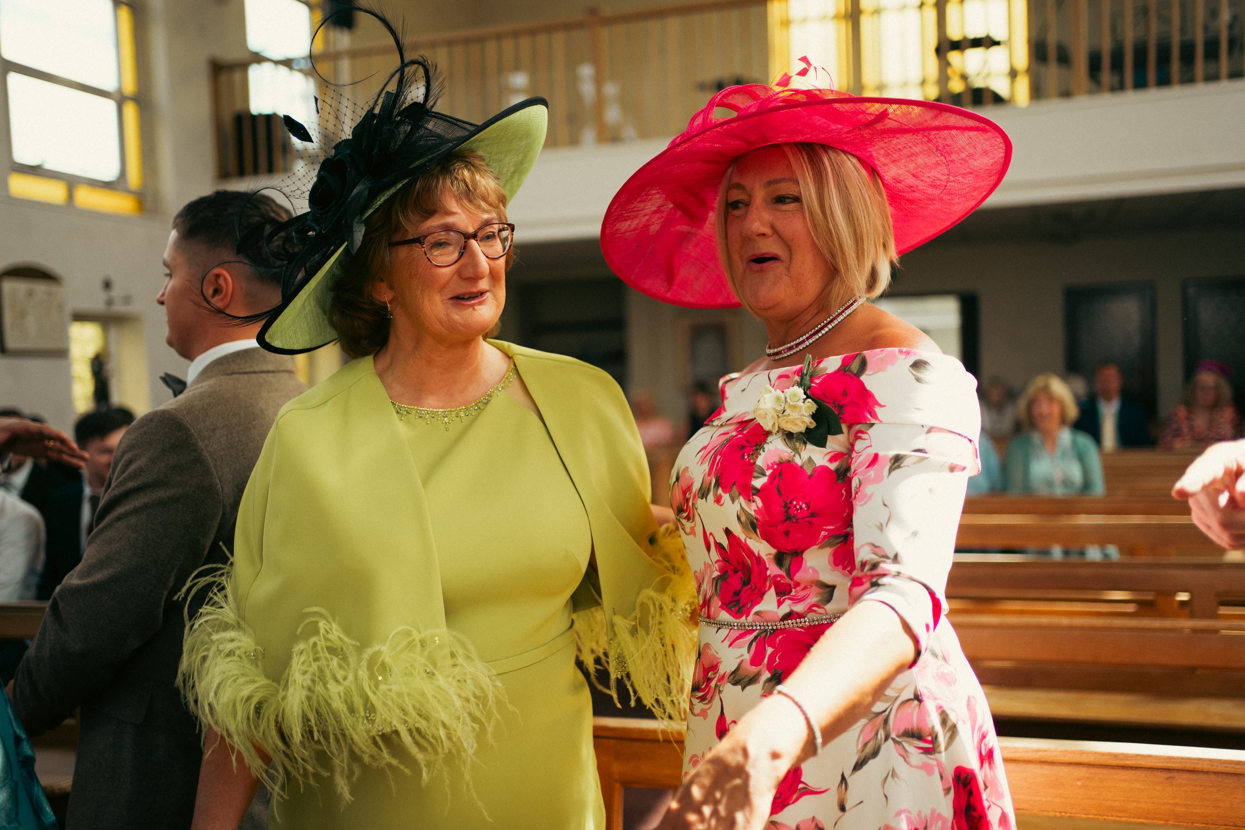 Two women wearing colorful dresses and large hats talking to each other inside a church during a wedding ceremony. The woman on the left is wearing a lime green dress with feather details and a matching hat with black embellishments. The woman on the